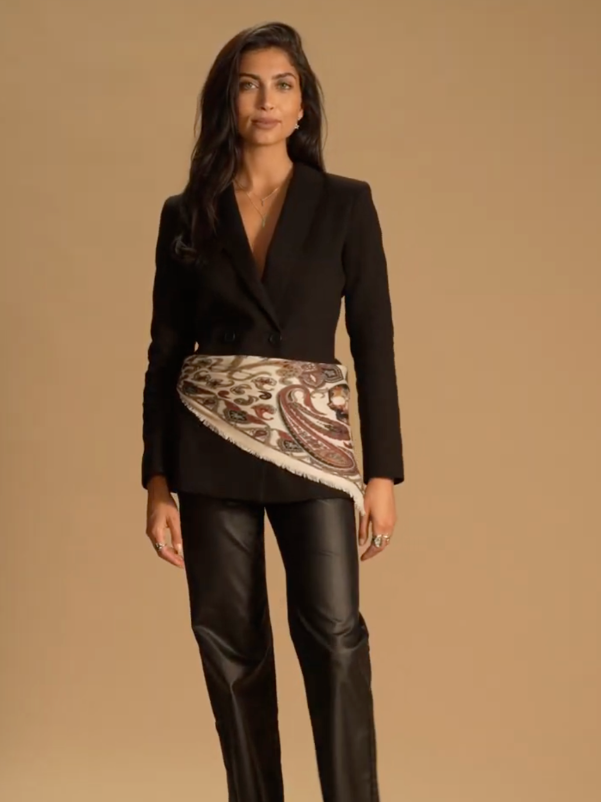 A model standing in front of a brown background wearing a black blazer with black leather trousers and a paisley silk scarf wrapped around her waist.