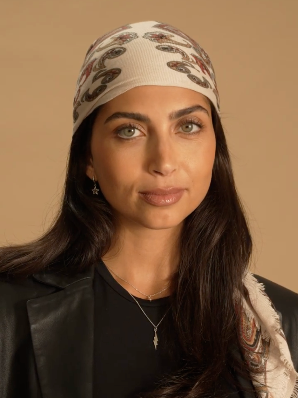 A close up shot of a model standing in front of a brown background wearing a black leather blazer over a black top. On her head she wears a paisley wool scarf tied as a bandana.