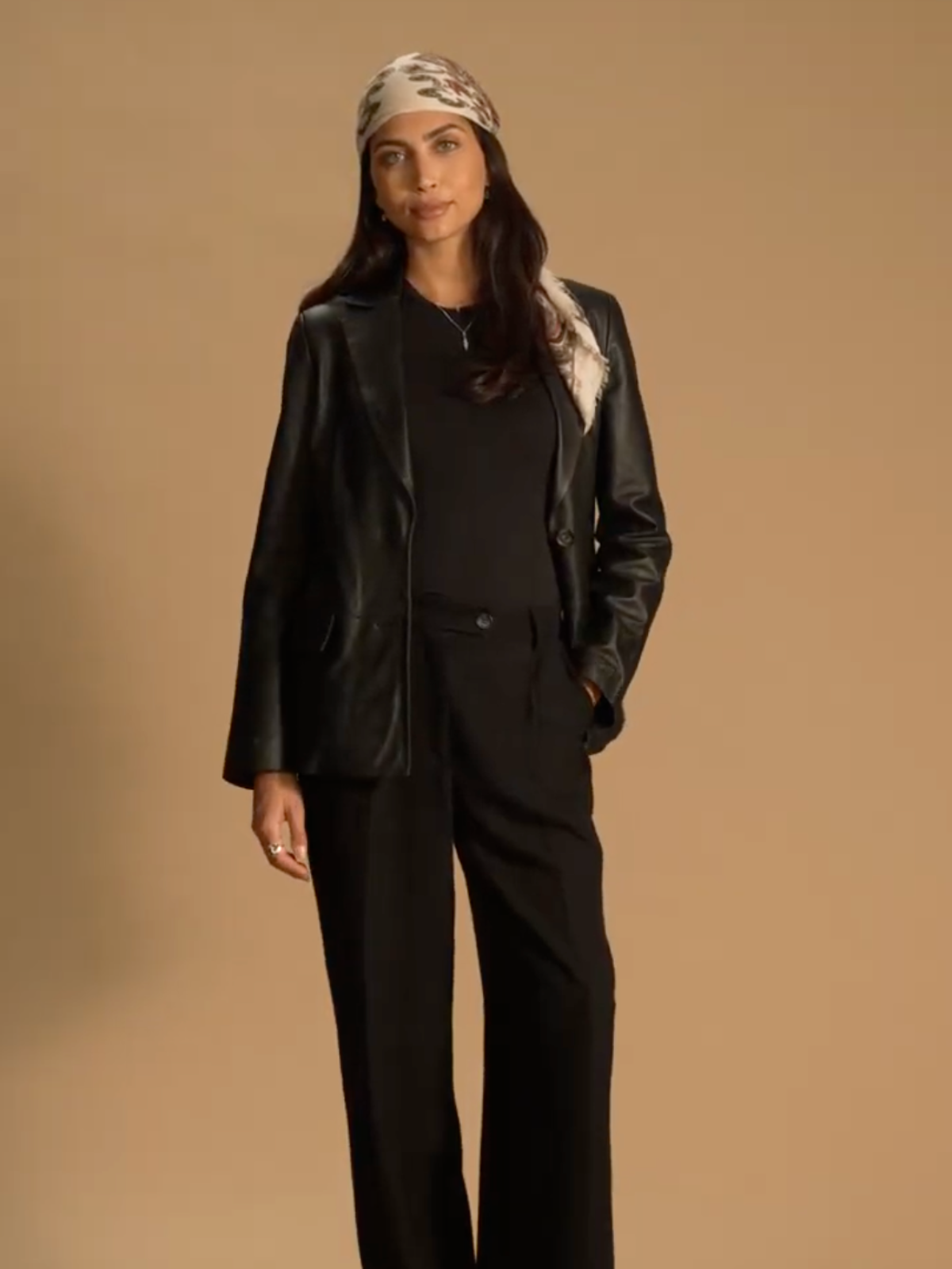 A model standing in front of a brown background wearing a Saint and Sofia leather blazer open over a black top with black trousers. On her head she wears a bandana made from a paisley print scarf.