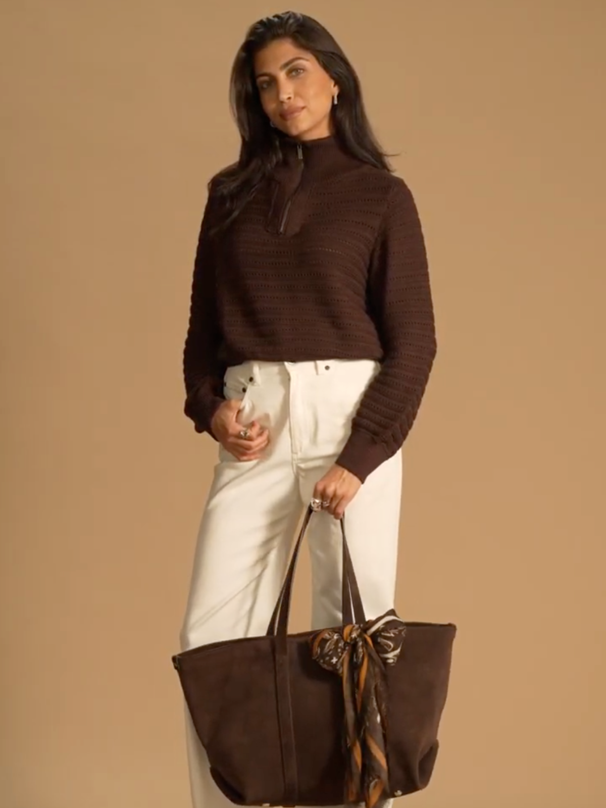 A model standing in front of a brown background wearing a brown knitted quarter zip jumper with white jeans. In her hand she holds a brown suede Saint and Sofia bag with the Wren paisley scarf in brown tied around the handle.
