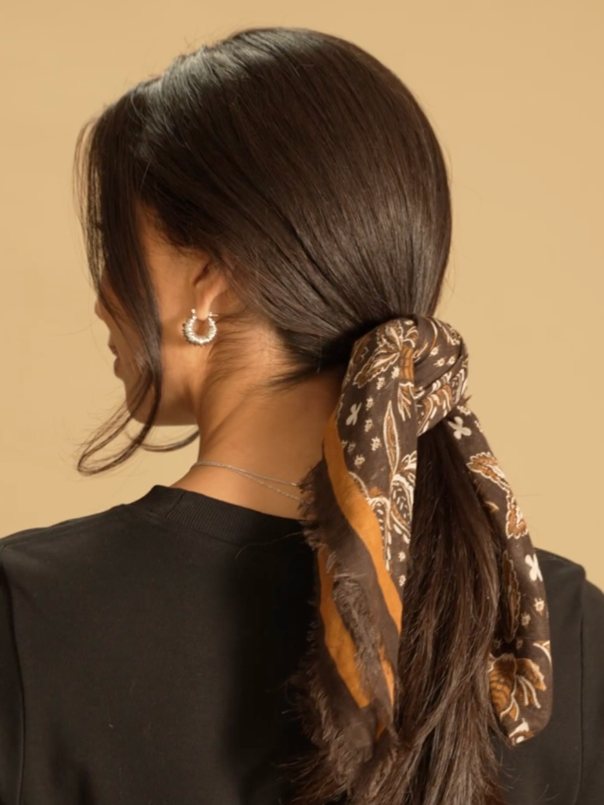A model standing with her back to the camera and her hair pulled back into a low ponytail. She wears a Saint and Sofia scarf wrapped around her hair.