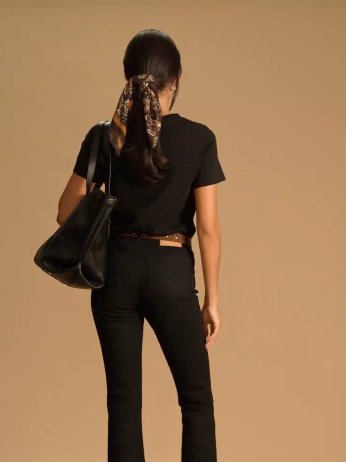 A model in front of a brown background wearing a black short sleeve top with black flared-leg jeans. She stands with her back to the camera with a black tote bag over her shoulder. Her hair is pulled back into a low pony and finished with a paisley scarf wrapped around the hair tie.