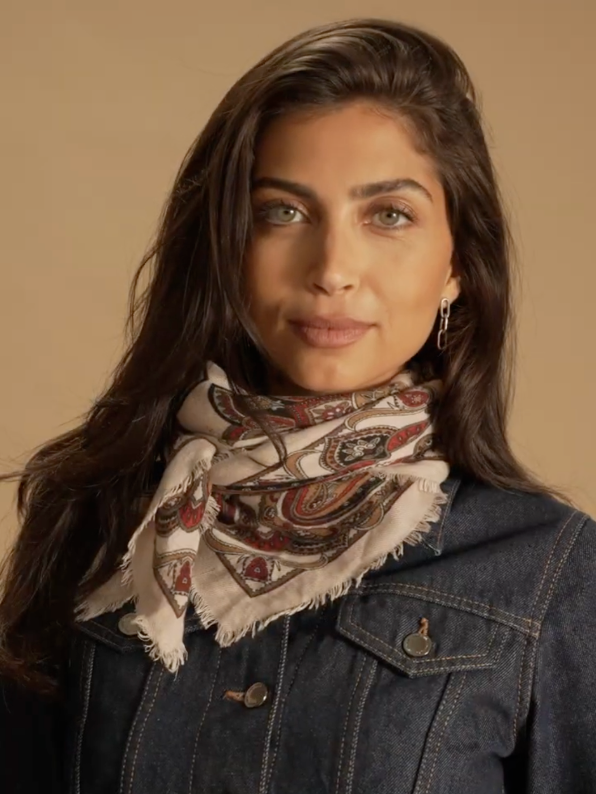 A model standing in front of a brown background wearing a denim jacket and a beige paisley neck tie. The shot is close up.