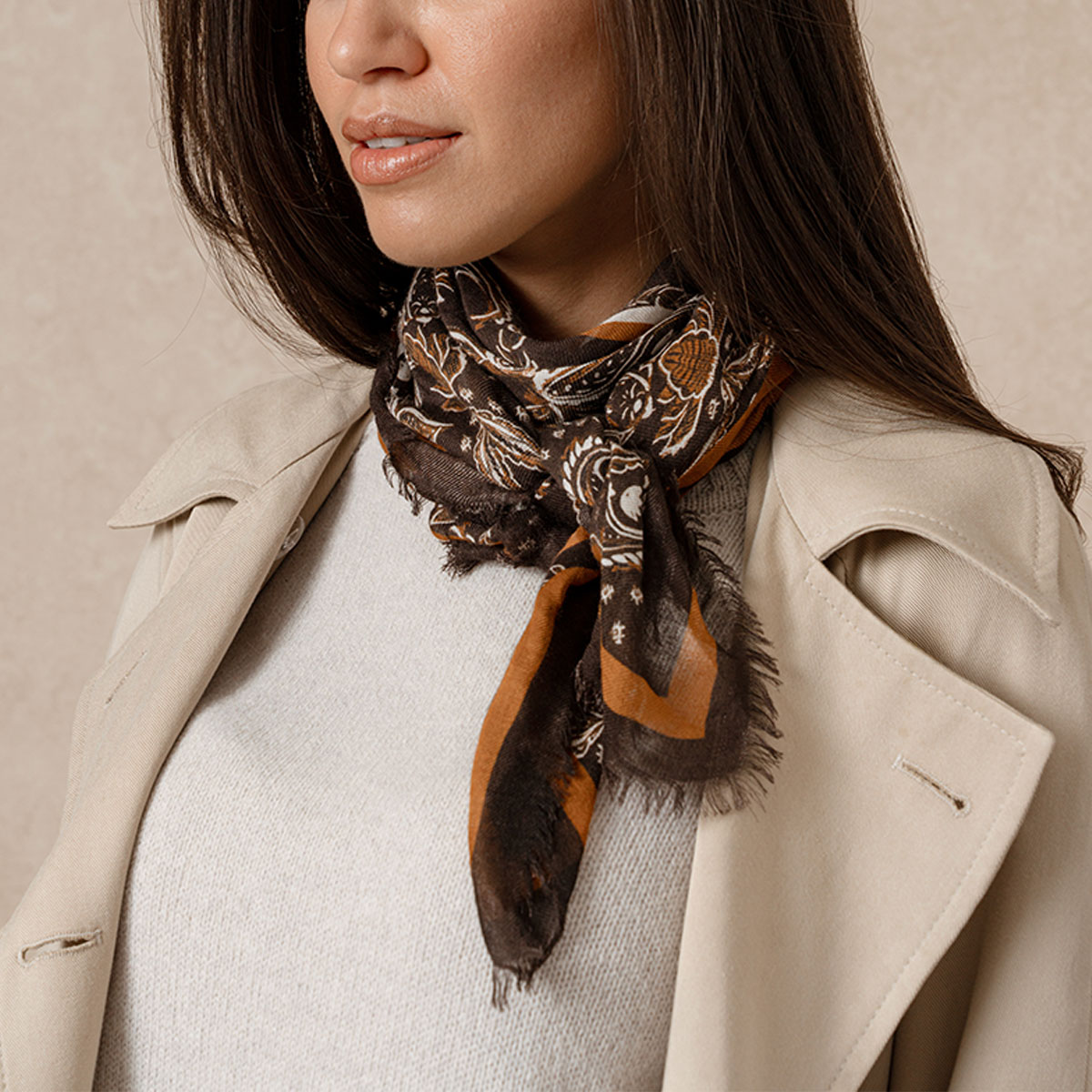 A model wearing a beige trench coat and cream jumper standing in front of a beige background. She wears the Saint and Sofia wren scarf around her neck.