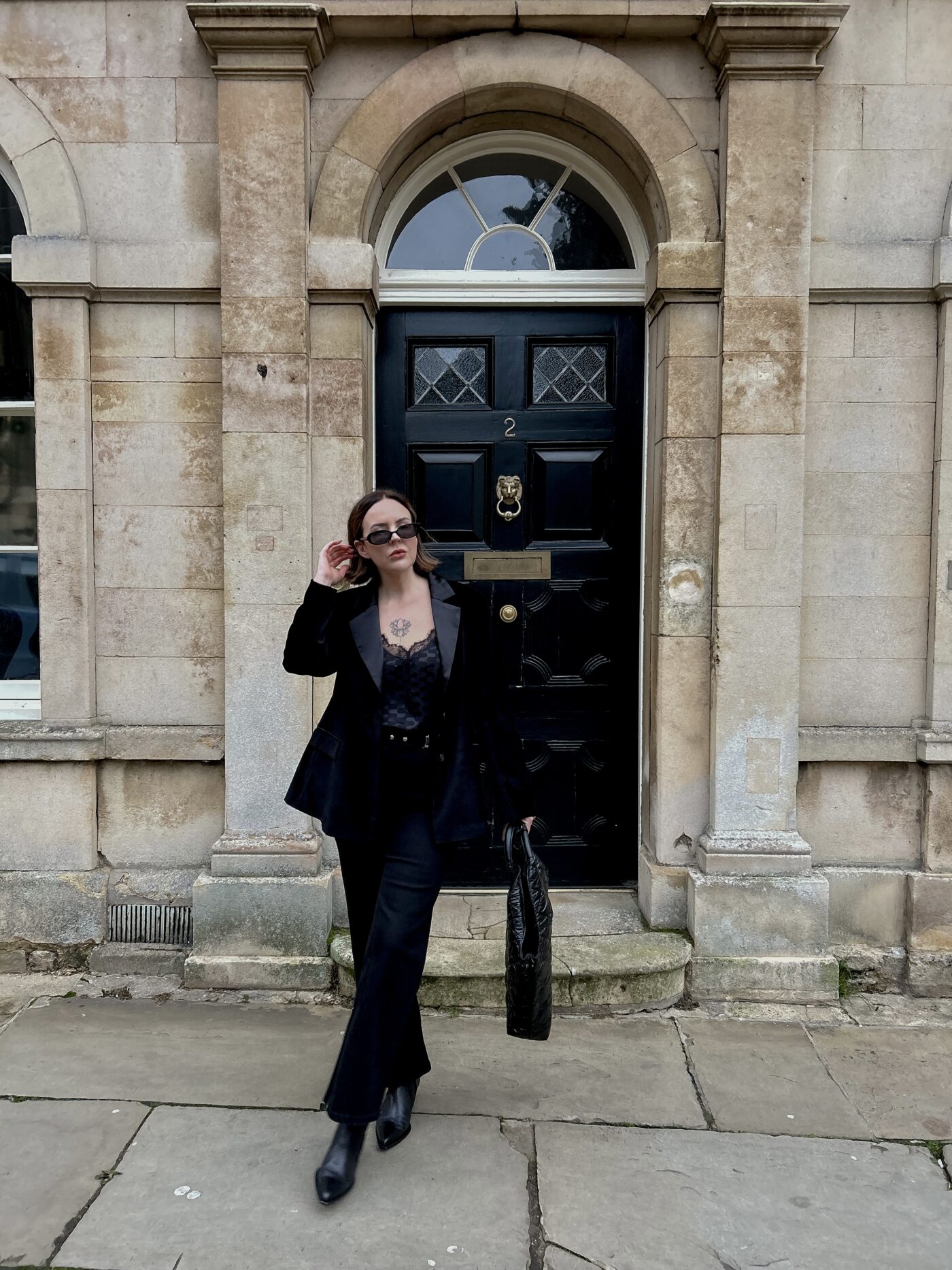 Leonie Holland stood outside a slab building wearing the Saint and Sofia Axel Single Breasted Blazer, a pair of black wide leg jeans, leather pointed toe boots, and the Soho quilted tote bag. All from the Saint and Sofia Black Friday sale.