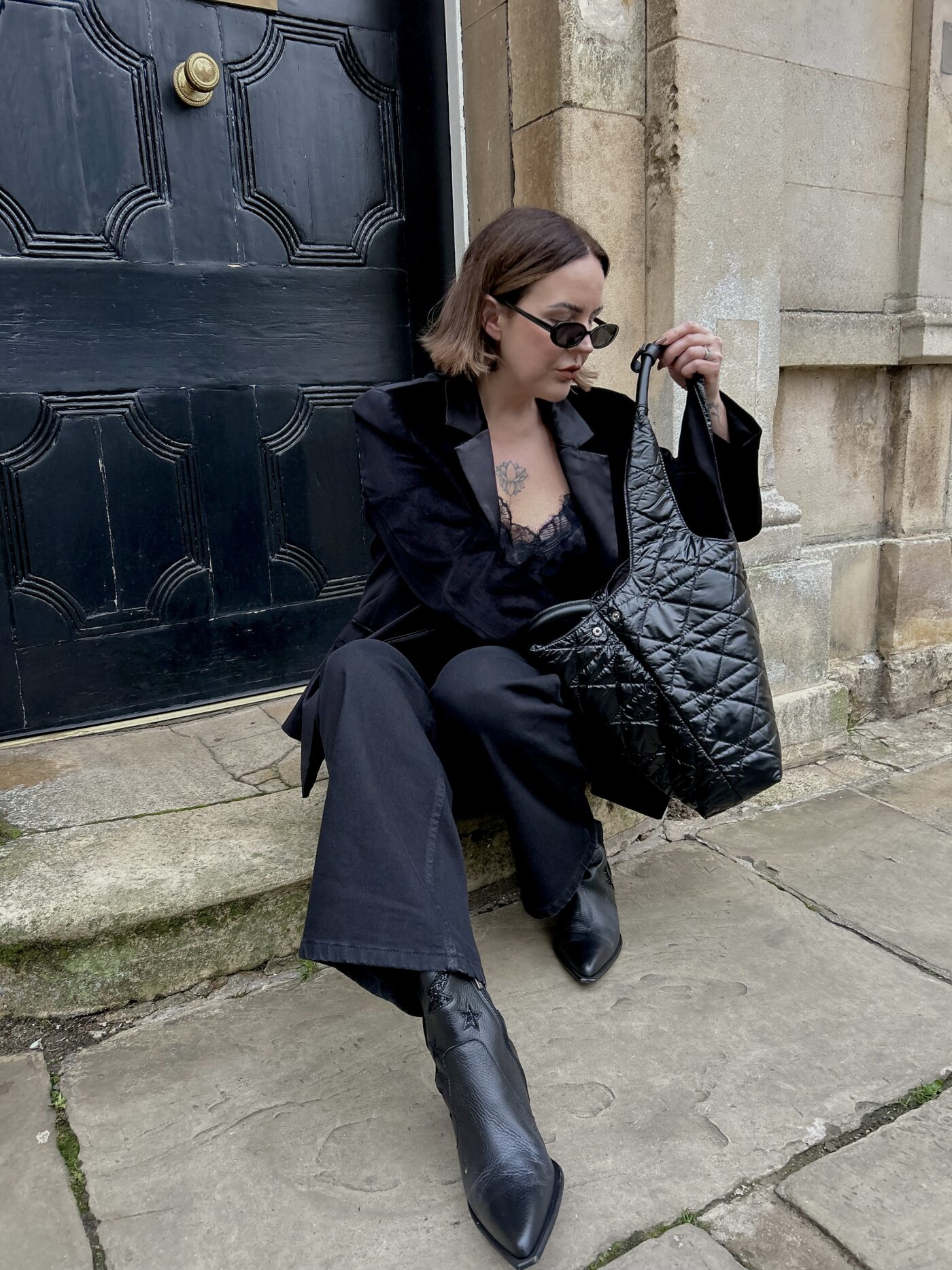 Leonie Holland sat on the doorstep outside a slab building wearing the Saint and Sofia Axel Single Breasted Blazer, a pair of black wide leg jeans, leather pointed toe boots, and the Soho quilted tote bag. All from the Saint and Sofia Black Friday sale.