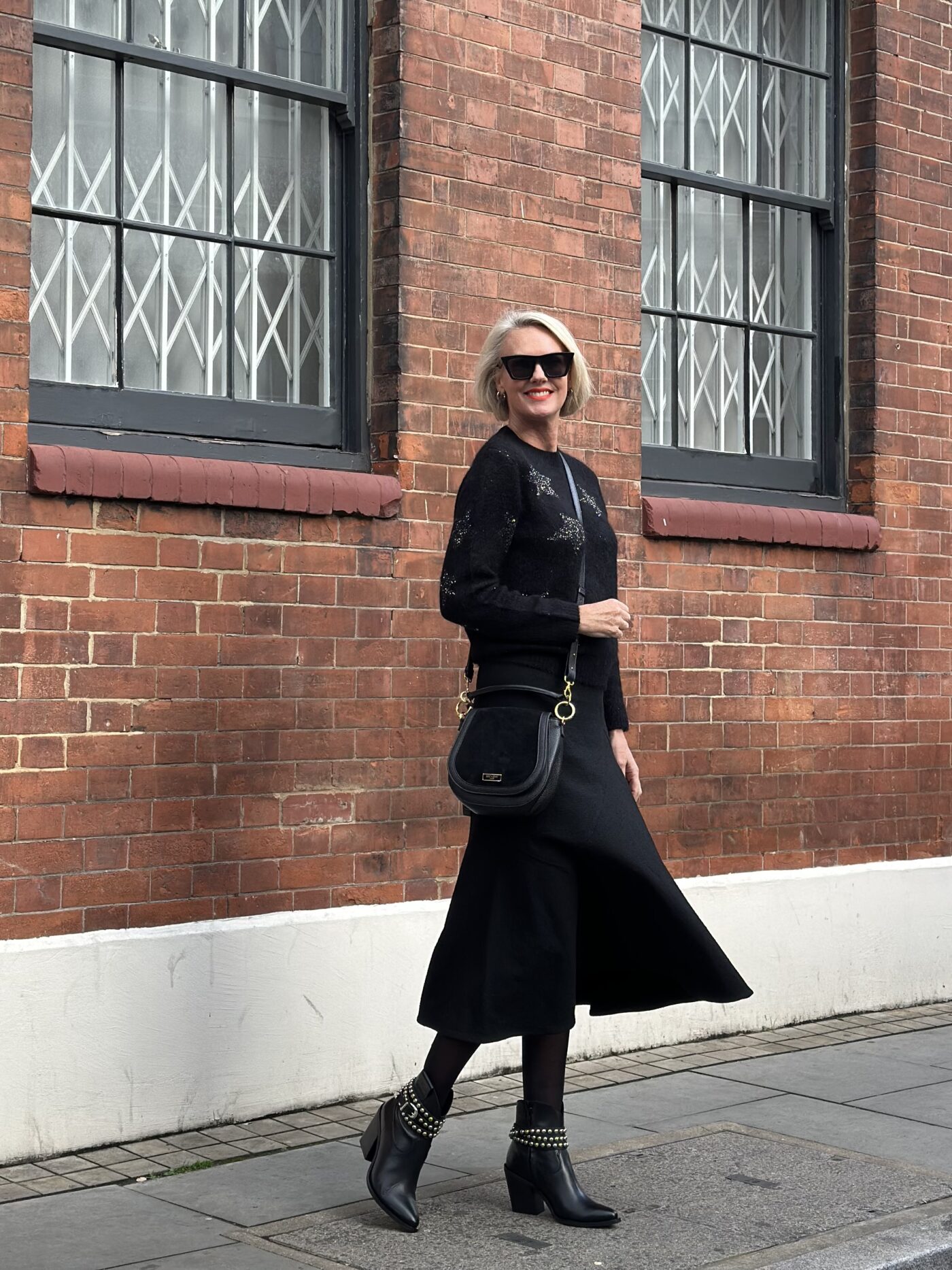 Coral Manson stood in front of a brick wall wearing the Saint and Sofia Indy Star Jumper with black sequin stars. She pairs it with a black midi skirt, leather boots, and a leather cross-body bag all from the Saint and Sofia Black Friday sale.
