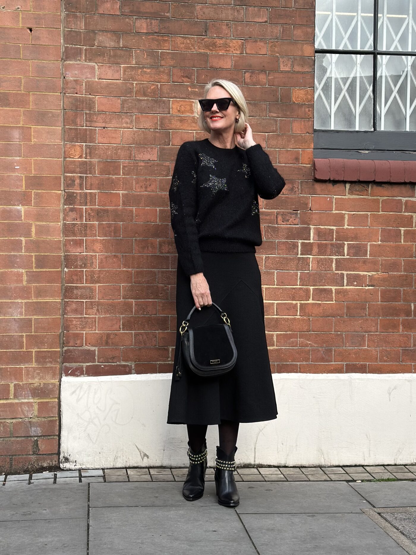 Coral Manson stood in front of a brick wall wearing the Saint and Sofia Indy Star Jumper with black sequin stars. She pairs it with a black midi skirt, leather boots, and a leather cross-body bag all from the Saint and Sofia Black Friday sale.