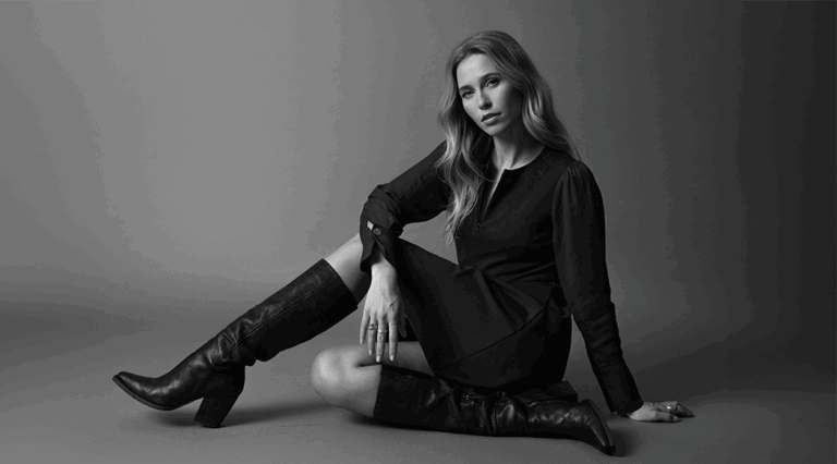A black and grey image of a model sat on the ground wearing a black long-sleeved midi dress with knee high leather boots.