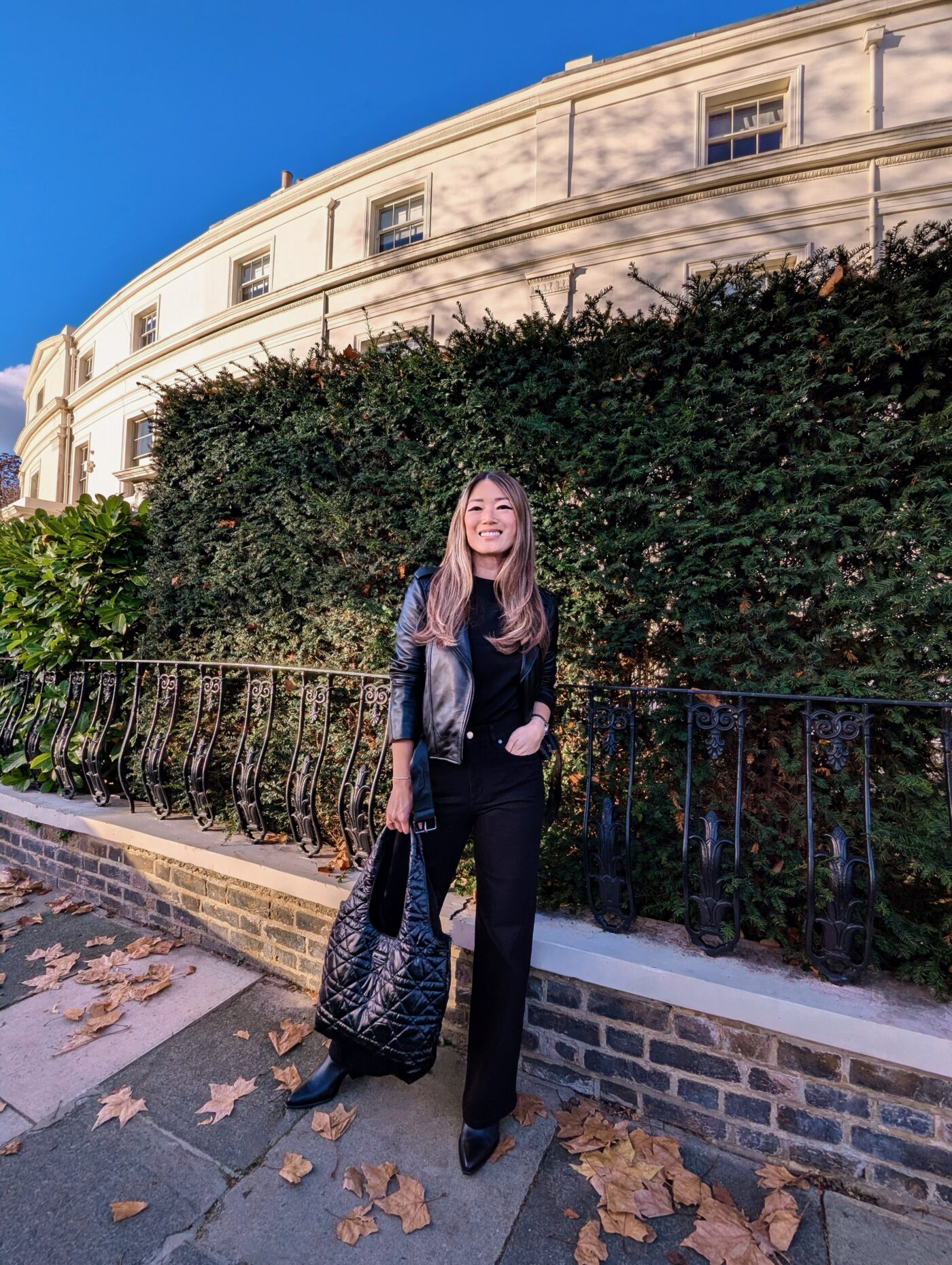 Sarah Highfield stood in front of a black metal fence wearing an all-black outfit by Saint and Sofia. She wears the leather Luther Jacket, a pair of black wide leg jeans, leather boots, and a quilted nylon bag.
