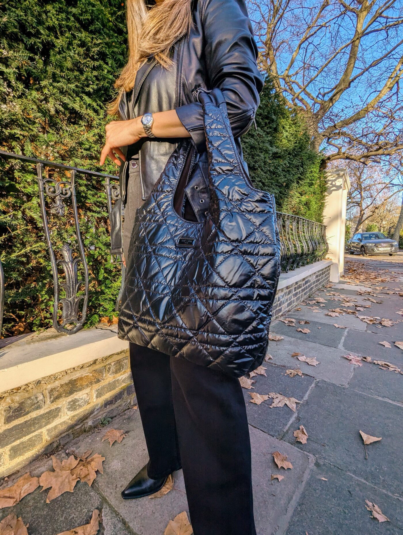 Sarah Highfield stood in front of a black metal fence wearing an all-black outfit by Saint and Sofia. She wears the leather Luther Jacket, a pair of black wide leg jeans, leather boots, and a quilted nylon bag. This picture is taken up close to the bag.
