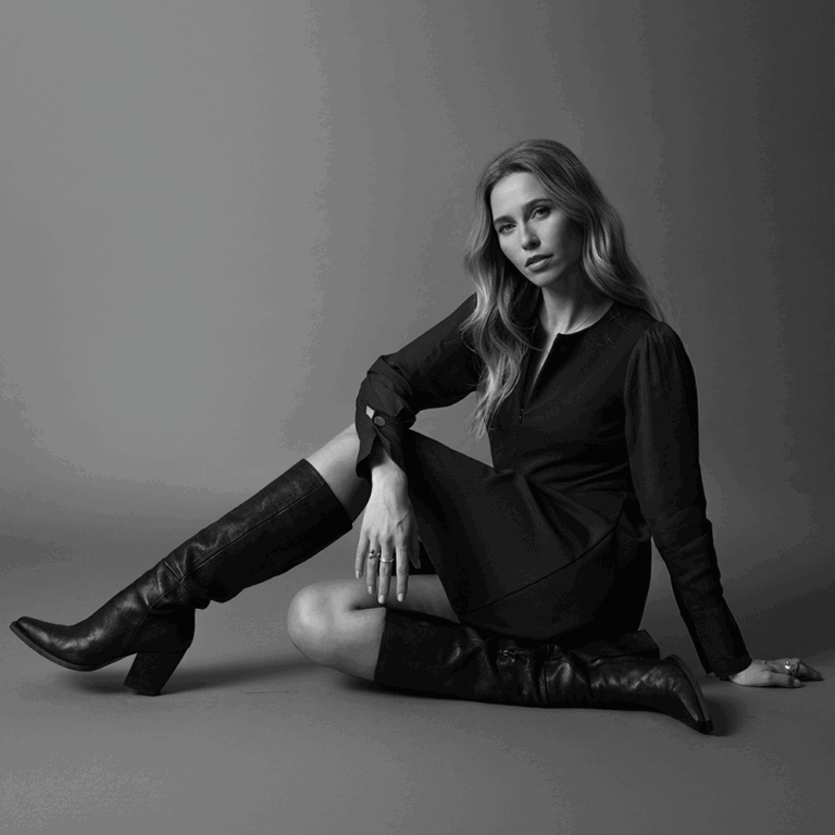A black and grey image of a model sat on the ground wearing a black long-sleeved midi dress with knee high leather boots.