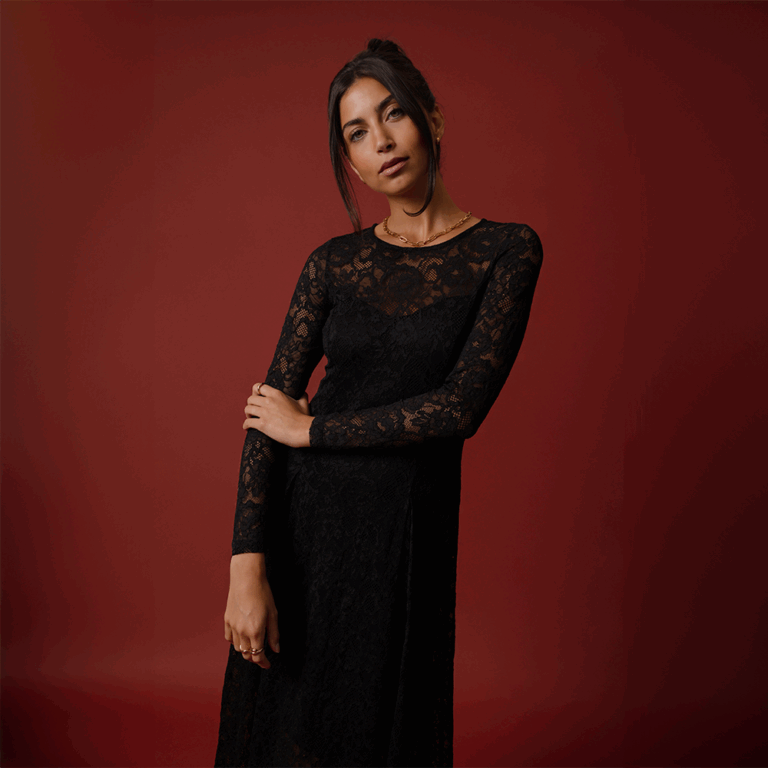 A model standing in front of a red background wearing a crew neck, long sleeved lace dress. A Christmas party dress.