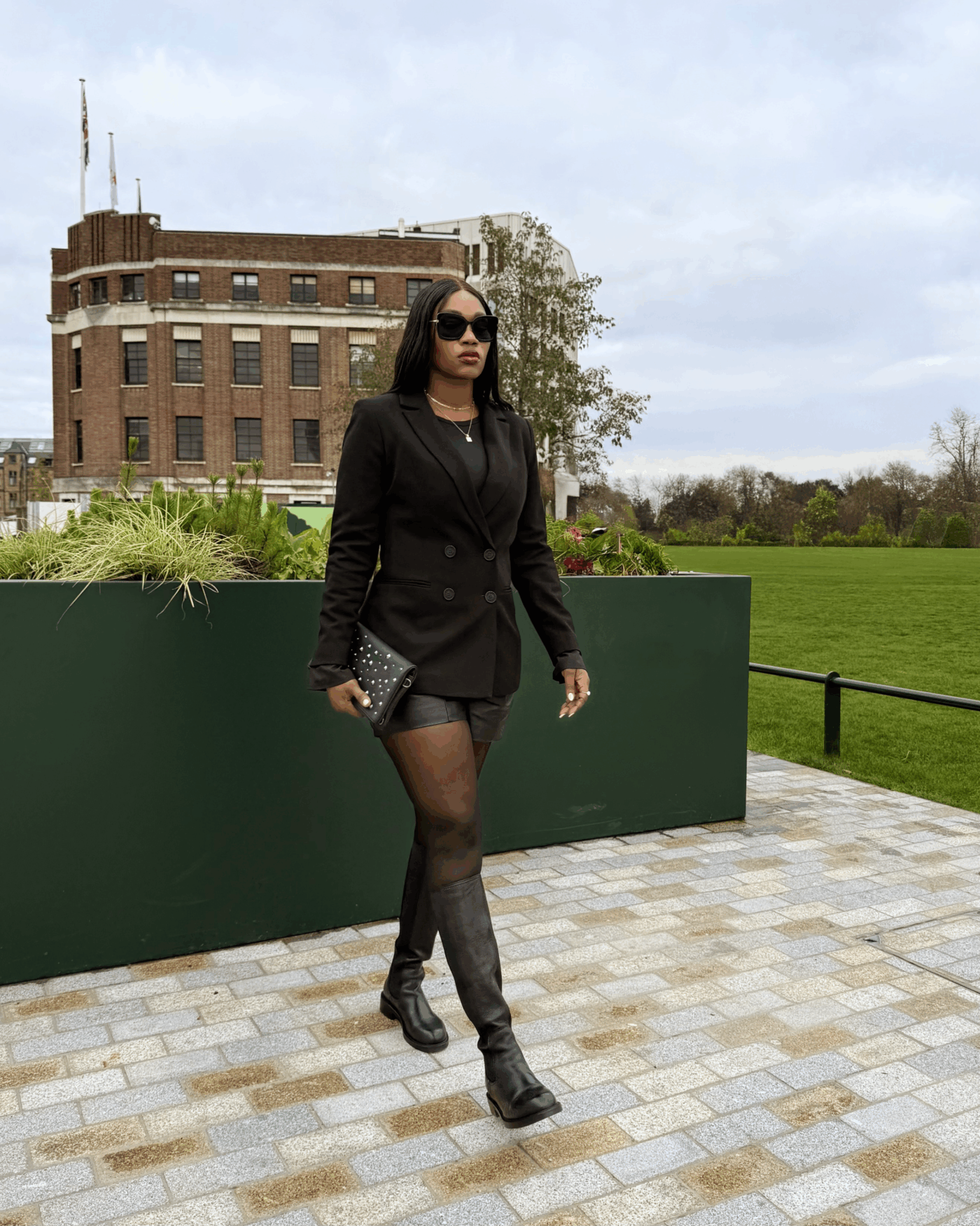 Titi Asu walking toward the camera outside. She wears the double-breasted Cambridge blazer by Saint and Sofia, fastened over a black tee and worn with black leather shorts, tights, and leather knee high boots. In her hand she holds a leather clutch decorated with gunmetal studs. All pieces are from the Saint and Sofia Black Friday sale.