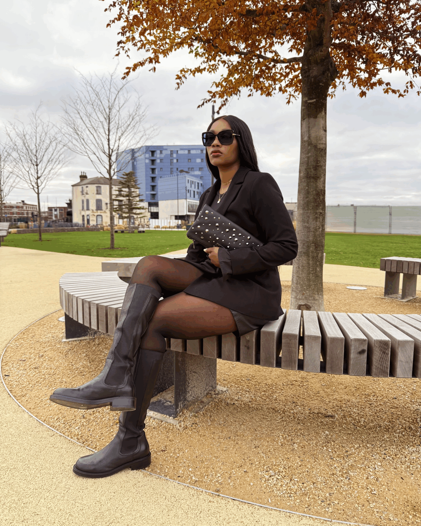 Titi Asu sat on a park bench. She wears the double-breasted Cambridge blazer by Saint and Sofia, fastened over a black tee and worn with black leather shorts, tights, and leather knee high boots. In her hand she holds a leather clutch decorated with gunmetal studs. All pieces are from the Saint and Sofia Black Friday sale.