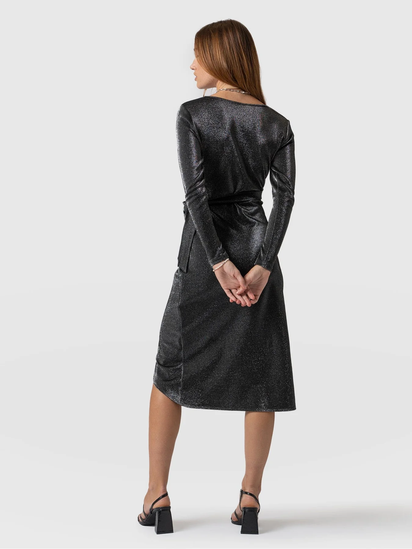 A model standing in front of a white background wearing the Amelia Wrap Dress in Silver by Saint and Sofia. A slim-fitting design with a square neckline, long sleeves, and a wrap-around finish with a knee-length skirt. In this image, she stands with her back to the camera.