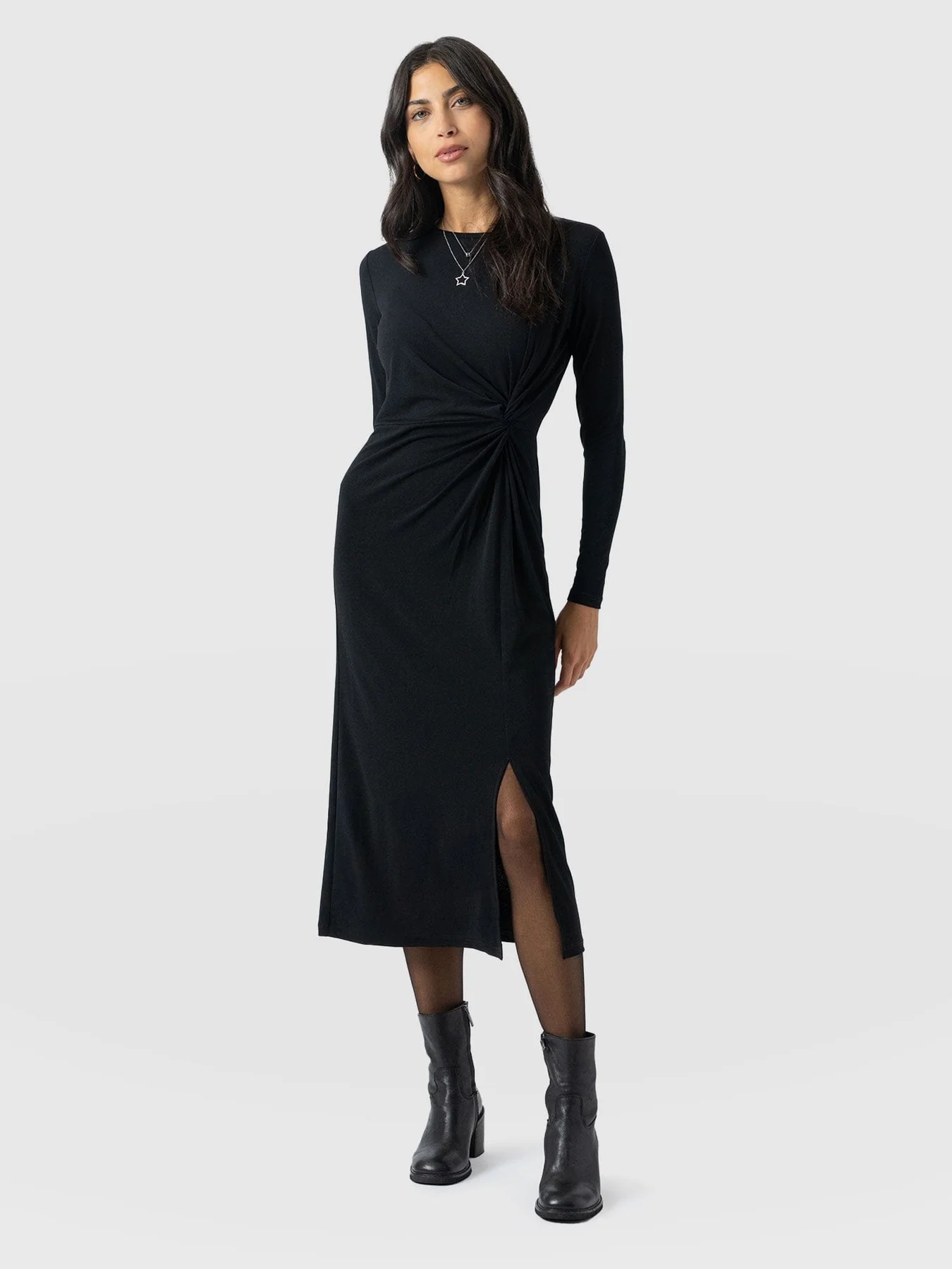 A model standing in front of a white background wearing the Bailey Knot Dress in Black. A slim-fitting crew neck dress with long sleeves, a midi length skirt, and a wrap-around aesthetic that finishes at a knot in the waist.
