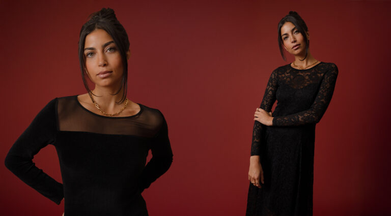 A rectangular image of two models stood in front of a red backdrop. The model on the left is standing closer to the camera wearing a black crew neck dress with a mesh panel at the decolletage. The model on the right is further away from the frame wearing a long sleeved lace midi dress.