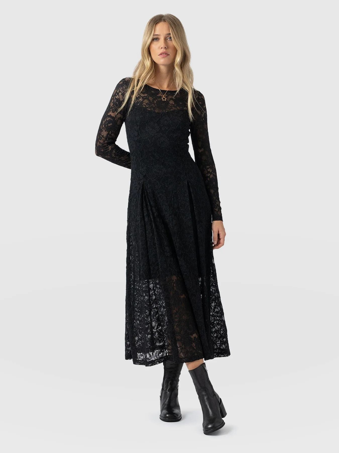 A model standing in front of a white background wearing the Lana Pleated Dress in Black Lace by Saint + Sofia. A crew neck design with long sleeves, and a full-bodied skirt. A lace overlay sits over a short black mini lining. A Christmas party dress.