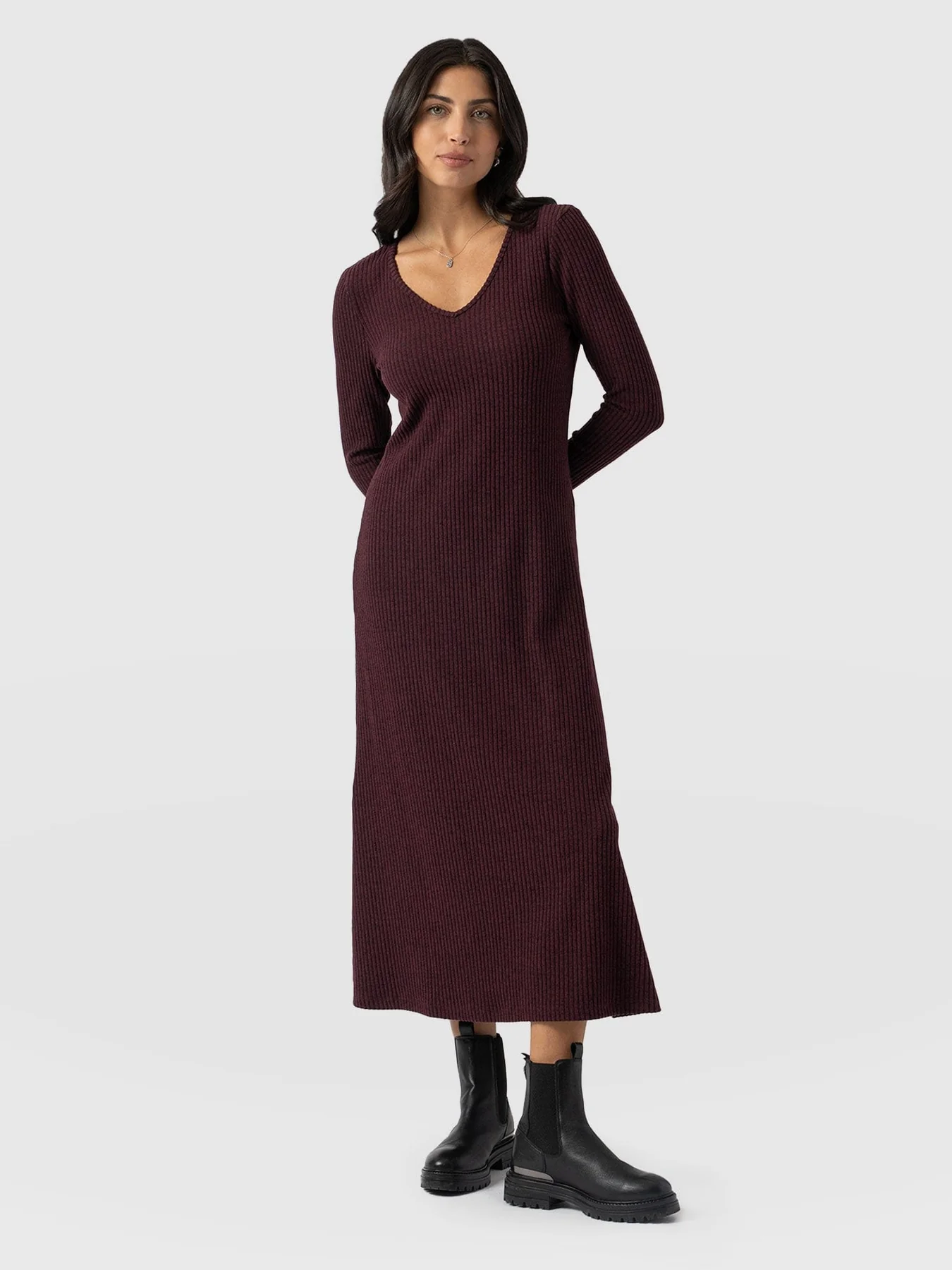 A model standing in front of a white background wearing the Sienna Dress in Burgundy by Saint and Sofia. A soft V neck dress handmade from a ribbed knit with long sleeves and a midi length skirt.