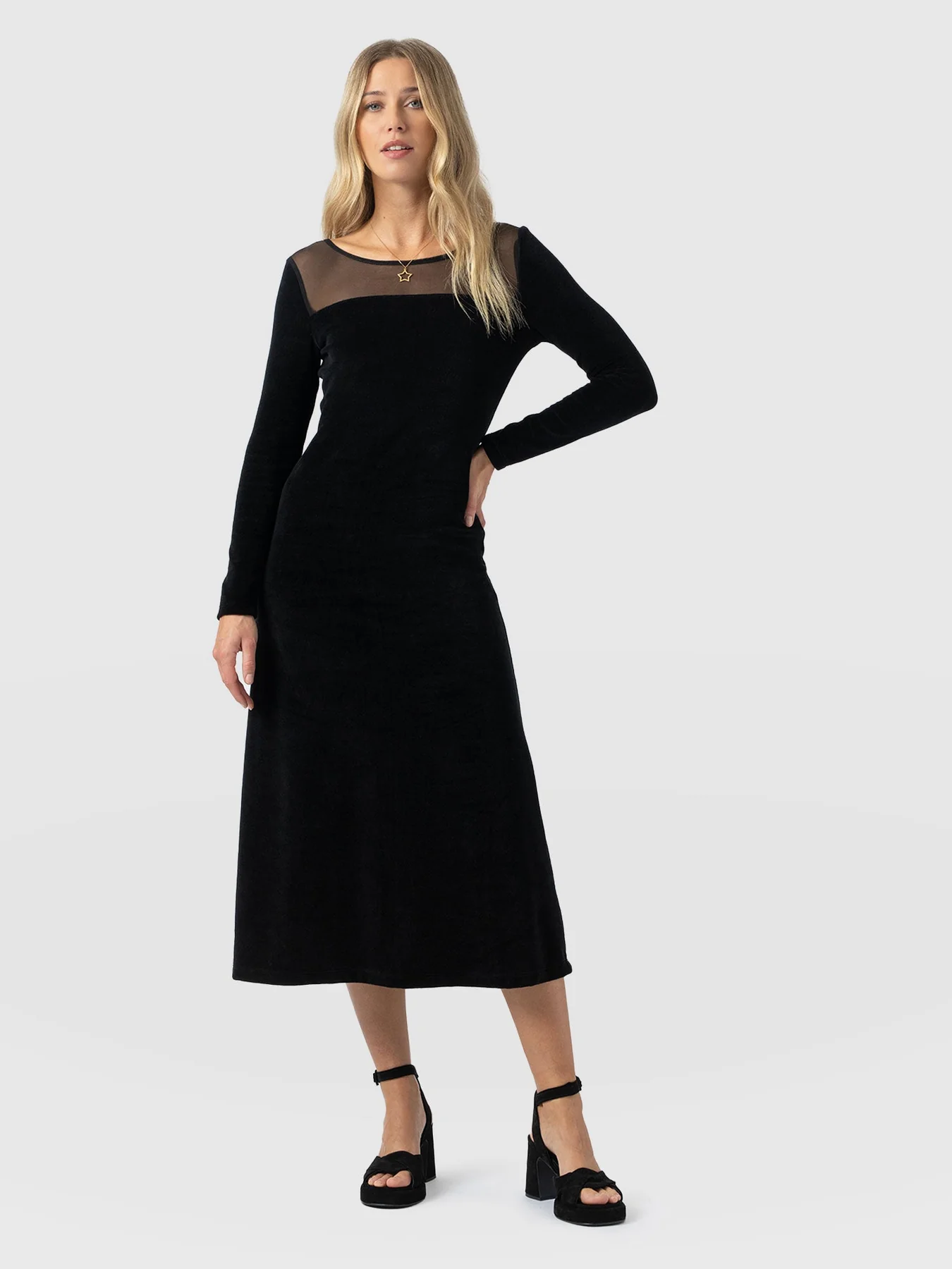 A model standing in front of a white background wearing the Storm Mesh Long Sleeve Dress in Black Velvet by Saint and Sofia. A crew neck black, slim-fitting dress with a mesh panel at the décolletage and long sleeves. A Christmas party dress.