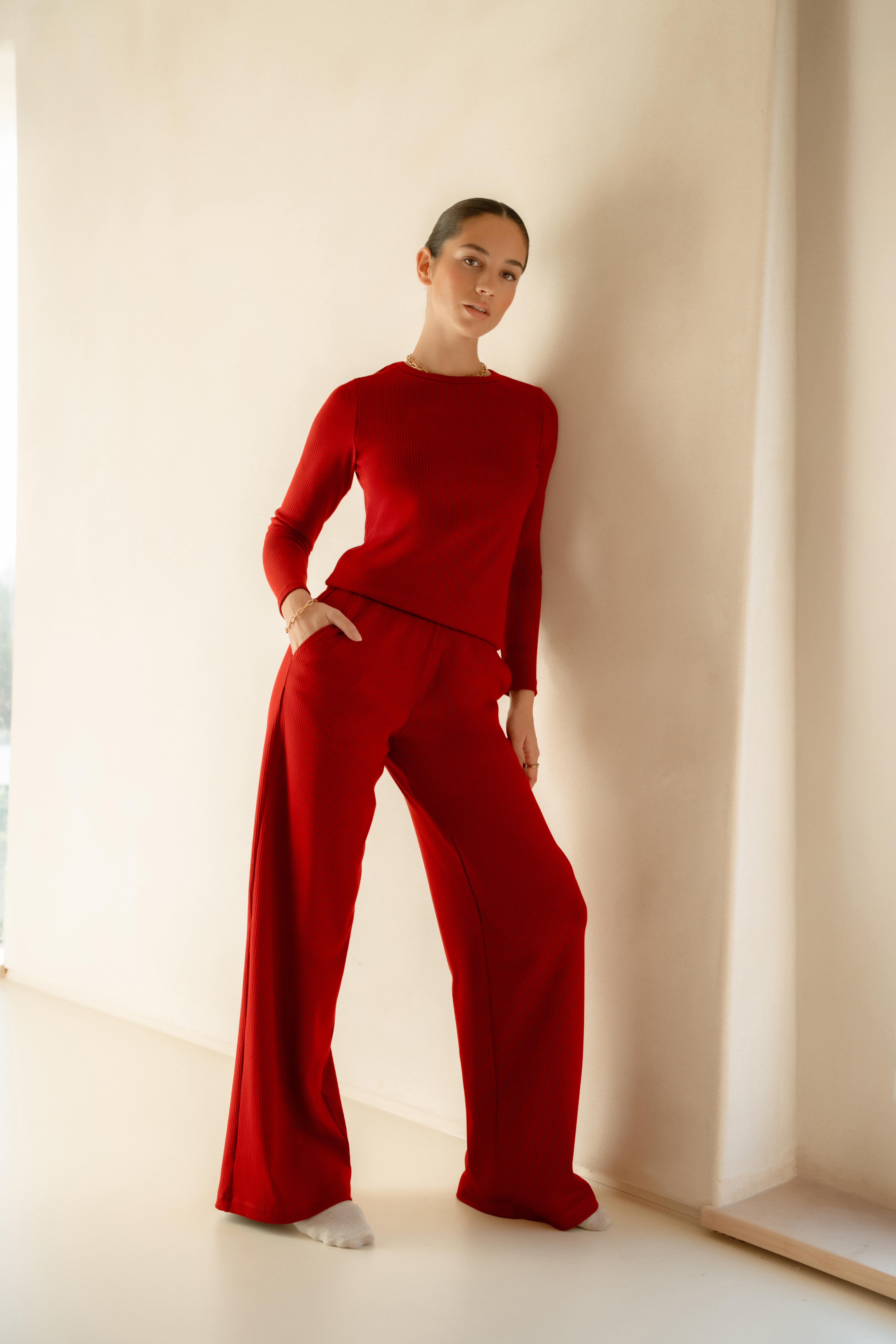 A model leaning against a wall wearing the Saint + Sofia red loungewear set. The set consists of a long sleeve crewneck ribbed top and wide legged elasticated ribbed trousers.