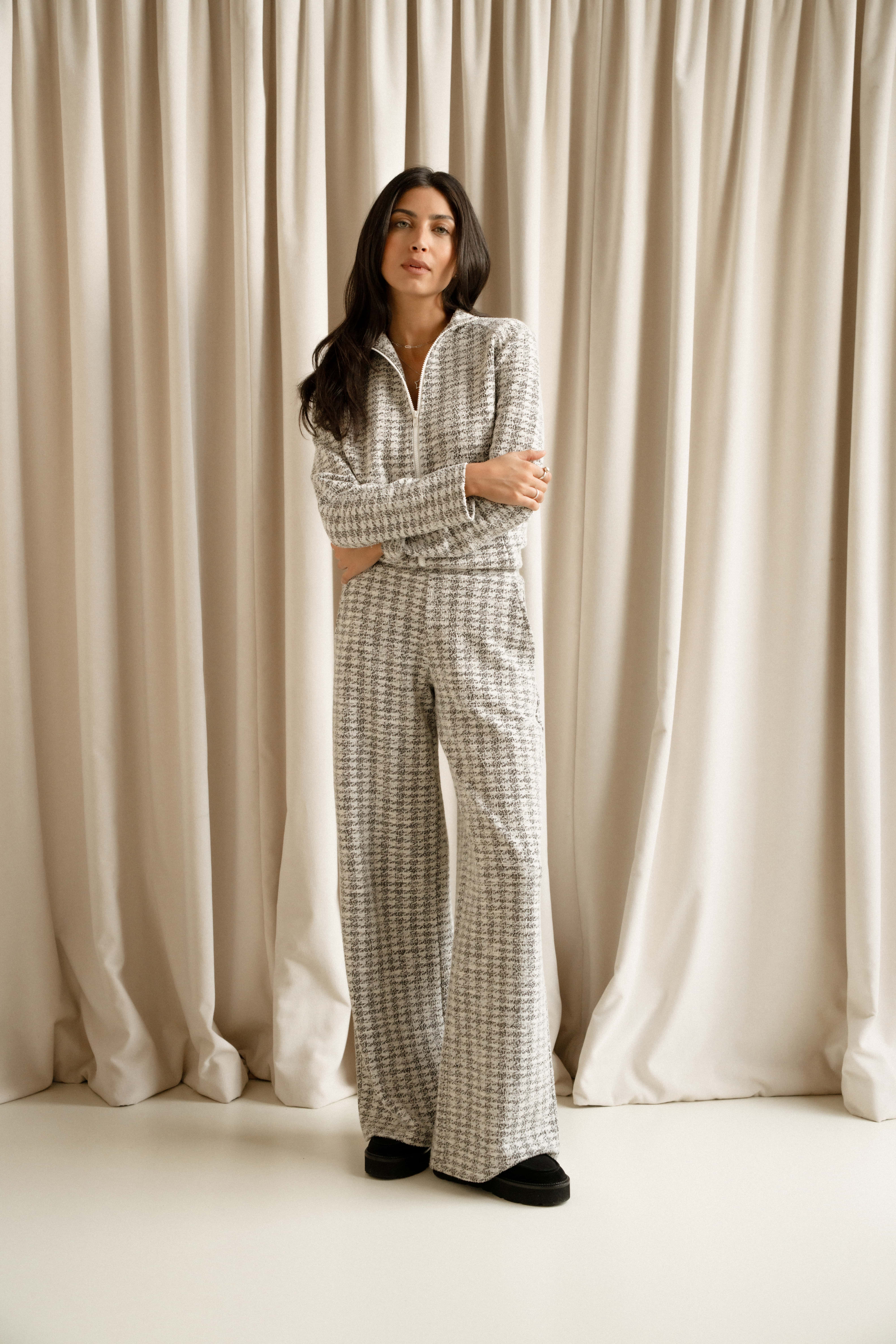 A model standing in front of a beige curtain wearing the Saint + Sofia boucle Keller Loungewear set. This set consists of a grey and white boucle zip up top and mathing wide leg trousers. The model wears the set with black slip on suede shoes.