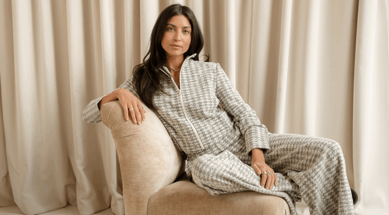 A model lounging on a cream sofa in front of a cream curtain back drop wearing the new Saint + Sofia grey and white boucle loungewear set.
