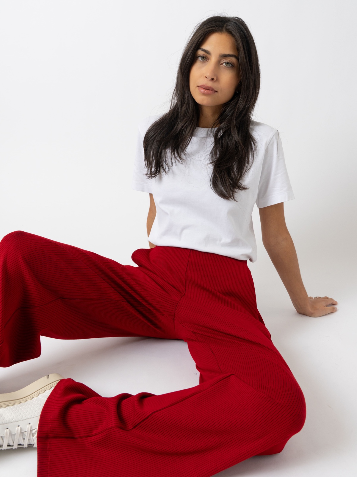 A model sat against a white backdrop wearing a white crew neck, short sleeve t-shirt and red ribbed wide leg trousers with white trainers.