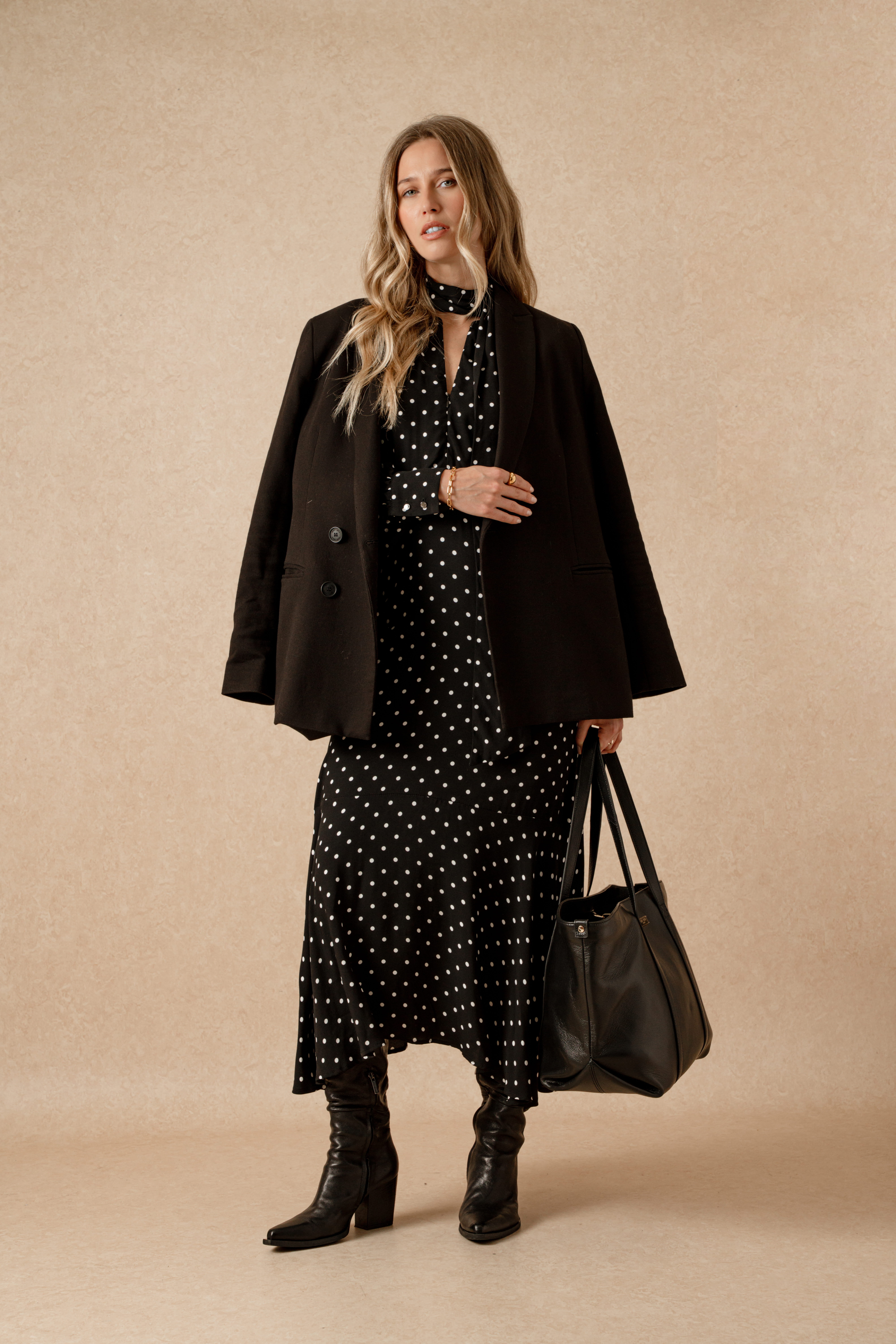 A model standing in front of a beige backdrop wearing the Saint and Sofia Adaline zip up dress. This black and white polka dot style features an adjustable zipped neckline with long sleeves, a silky neck tie, and midi length skirt. She wears it here under a black blazer with knee high leather boots and a black leather bag.