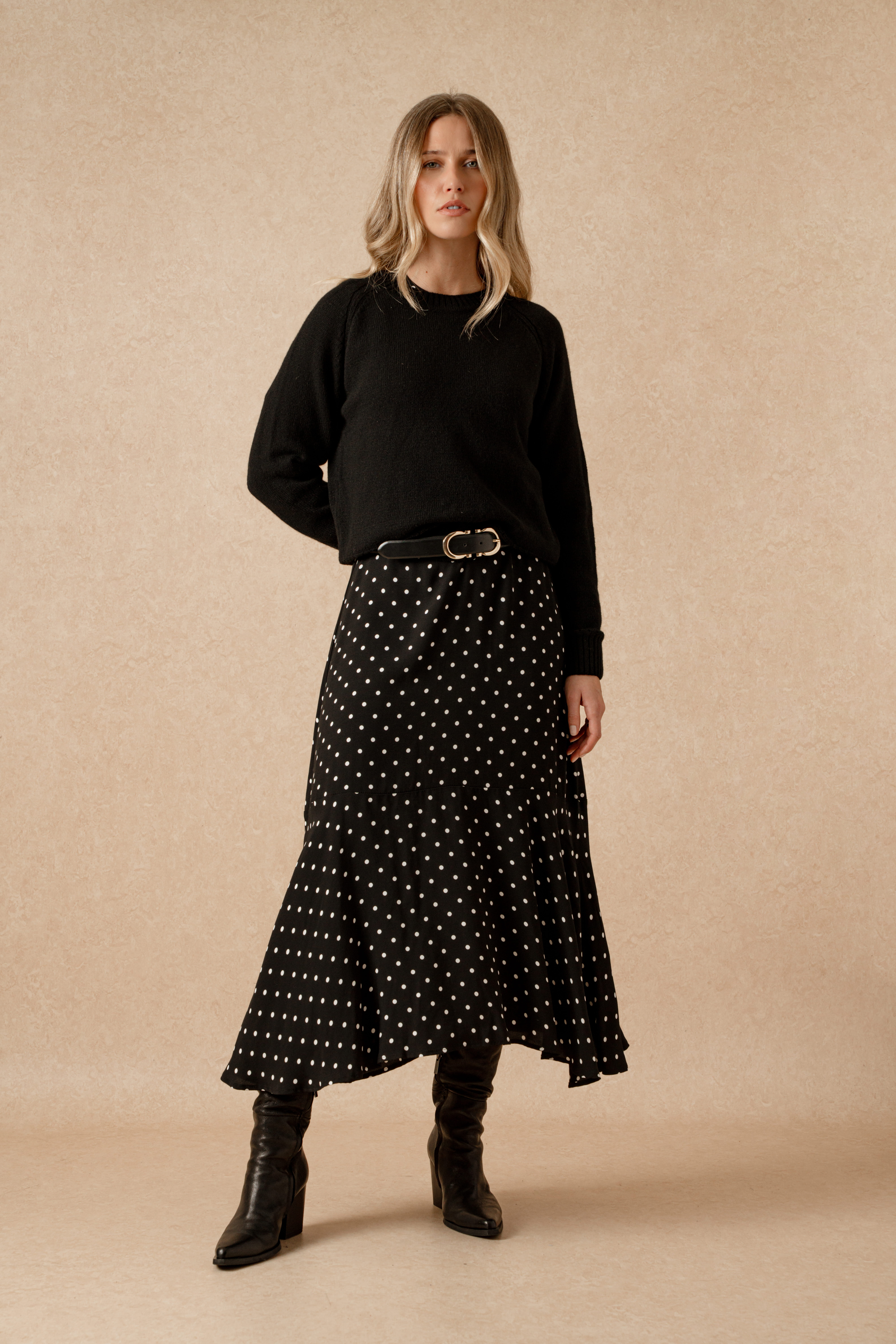 A model standing in front of a beige backdrop wearing the Saint and Sofia Adaline zip up dress. This black and white polka dot style features an adjustable zipped neckline with long sleeves, a silky neck tie, and midi length skirt. She wears it here with a black knitted jumper over the top to create the illusion of a skirt and jumper, and a leather belt around the waist with leather pointed toe boots.