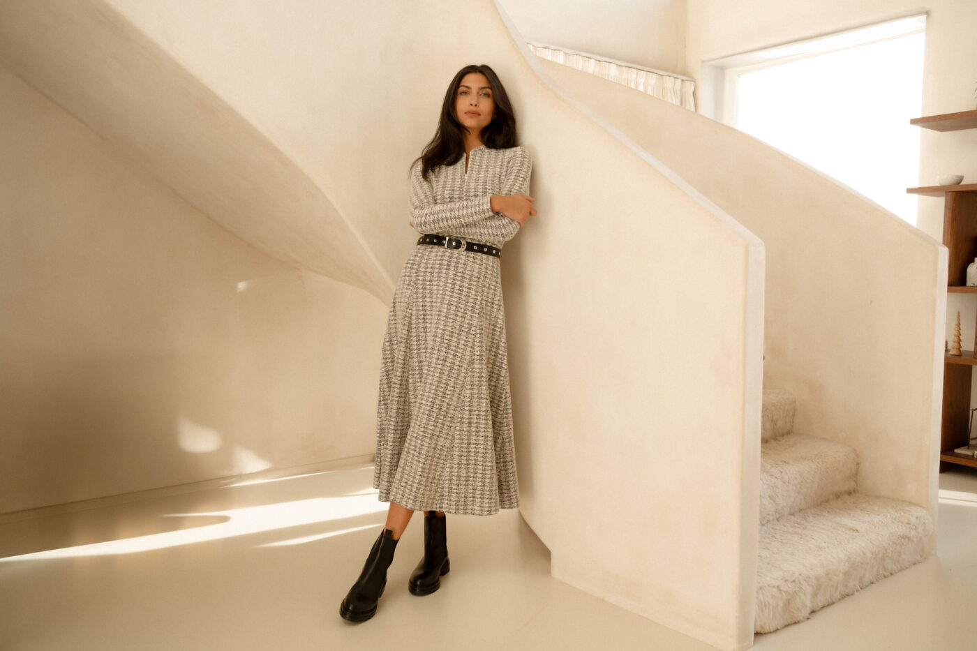 A model standing in front of a beige staircase wearing a black and white boucle-effect dress with long sleeves, a zipped front fastening, a black belt with silver metal studs, and black leather boots.