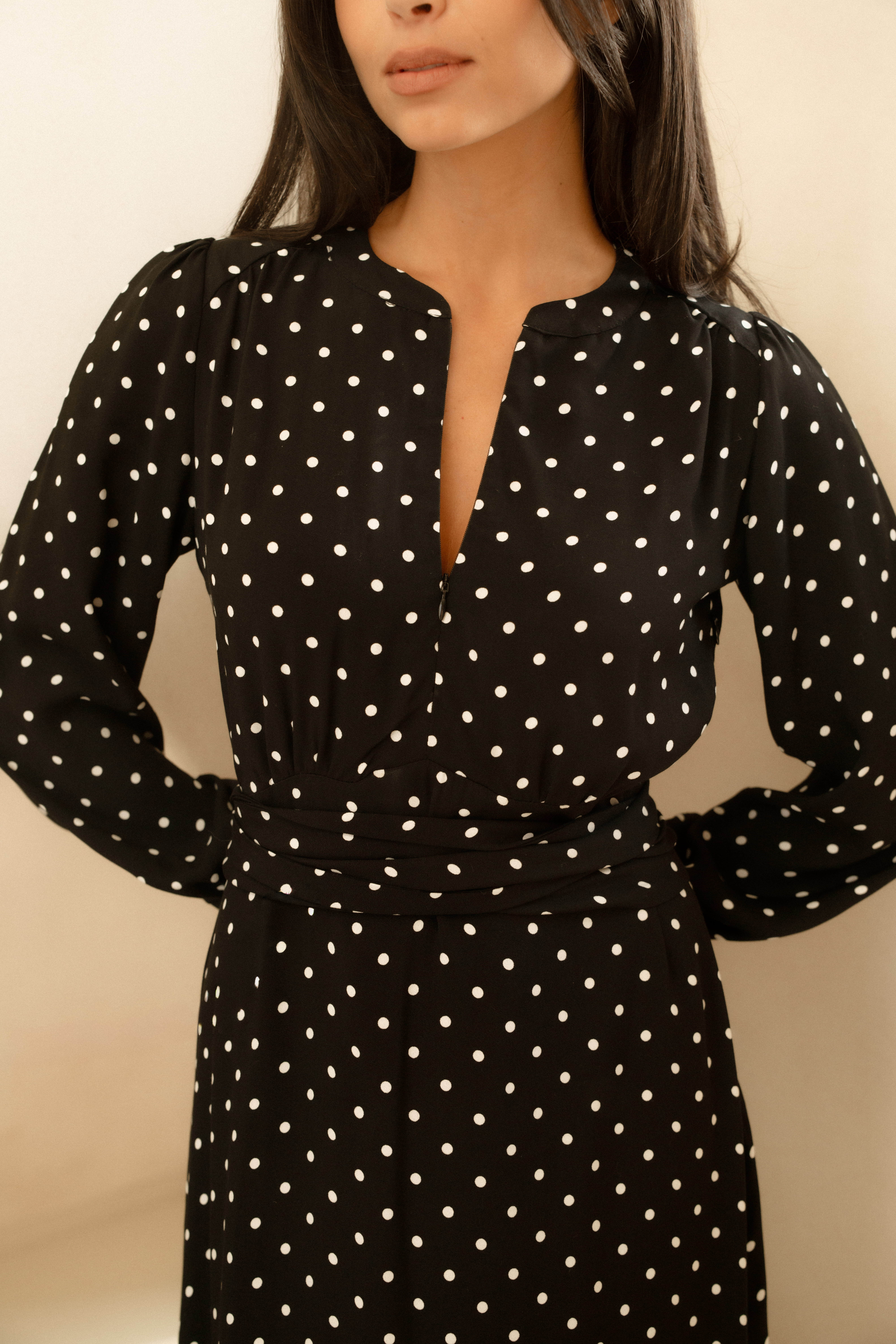 A close up image of a models torso as she wears a black and white polka dot dress with a V neckline, long sleeves, and cinched, belted waist.