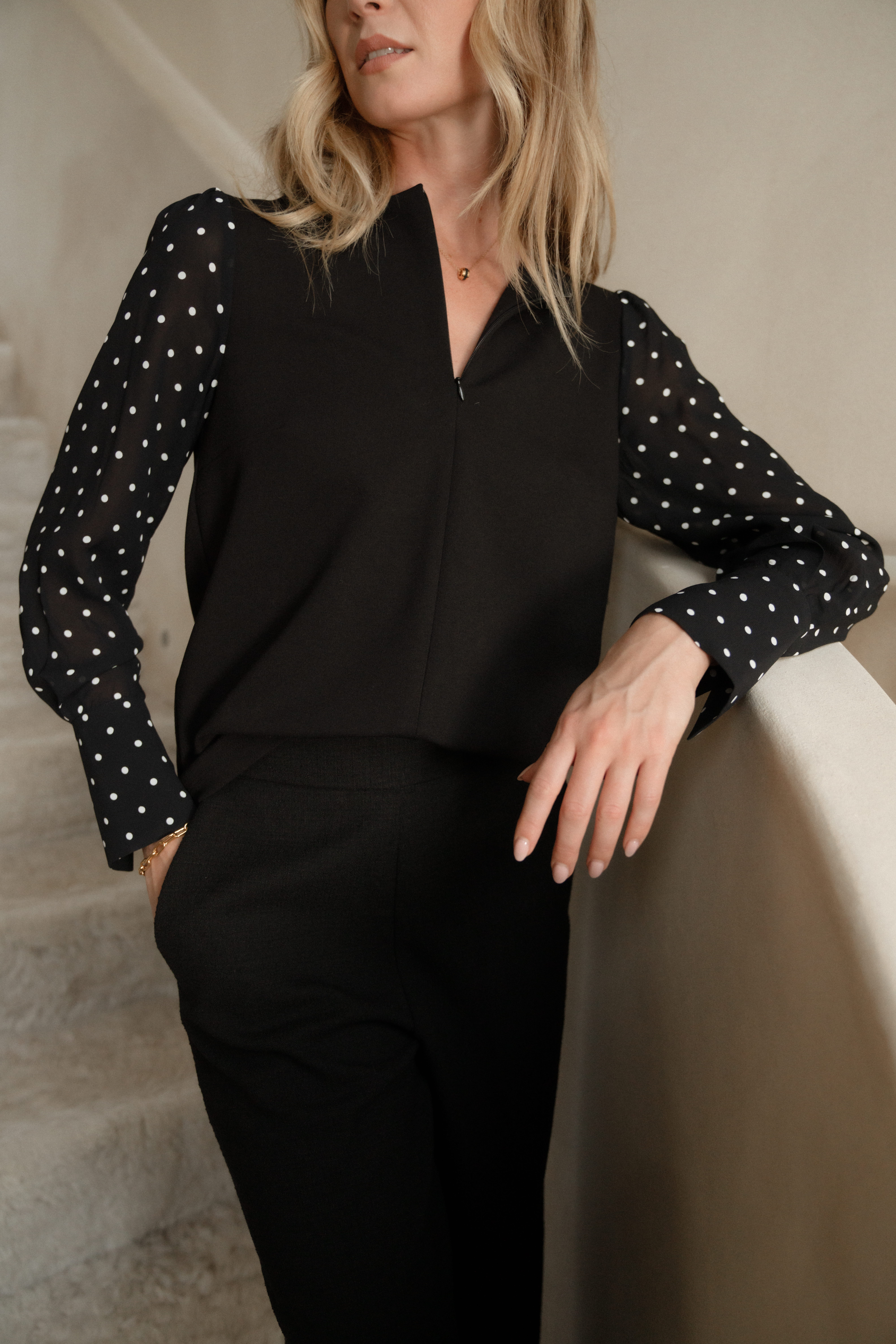A model wearing a black blouse with a zipped neckline and black and white polka dot sleeves. From Saint and Sofia's January collection. She wear this blouse with a pair of black wide leg trousers.
