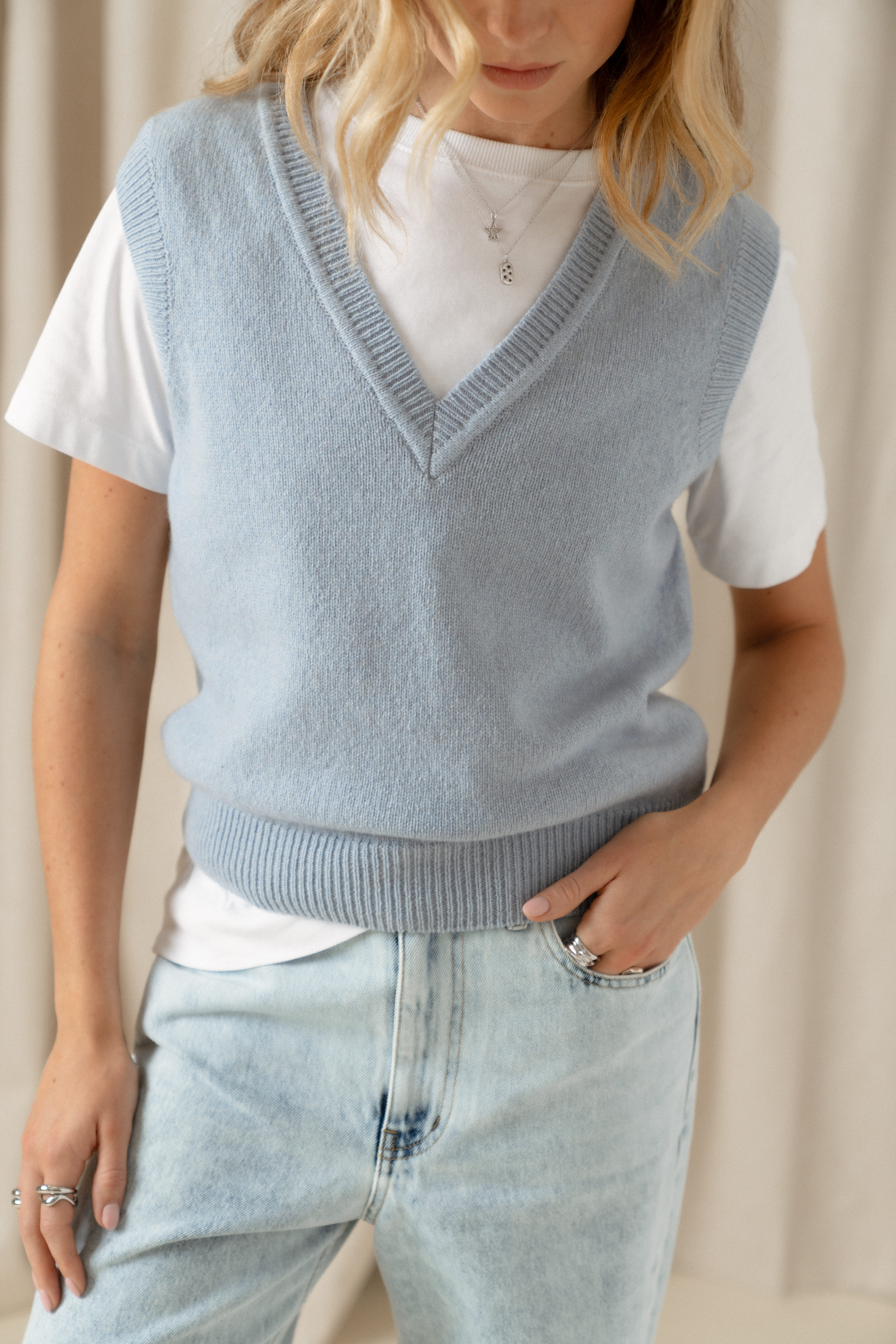 A shot of a model's torso as she wears a white short sleeve T-shirt with a crew neckline under a baby blue cashmere sweater vest by Saint and Sofia. Paired with light blue denim jeans.