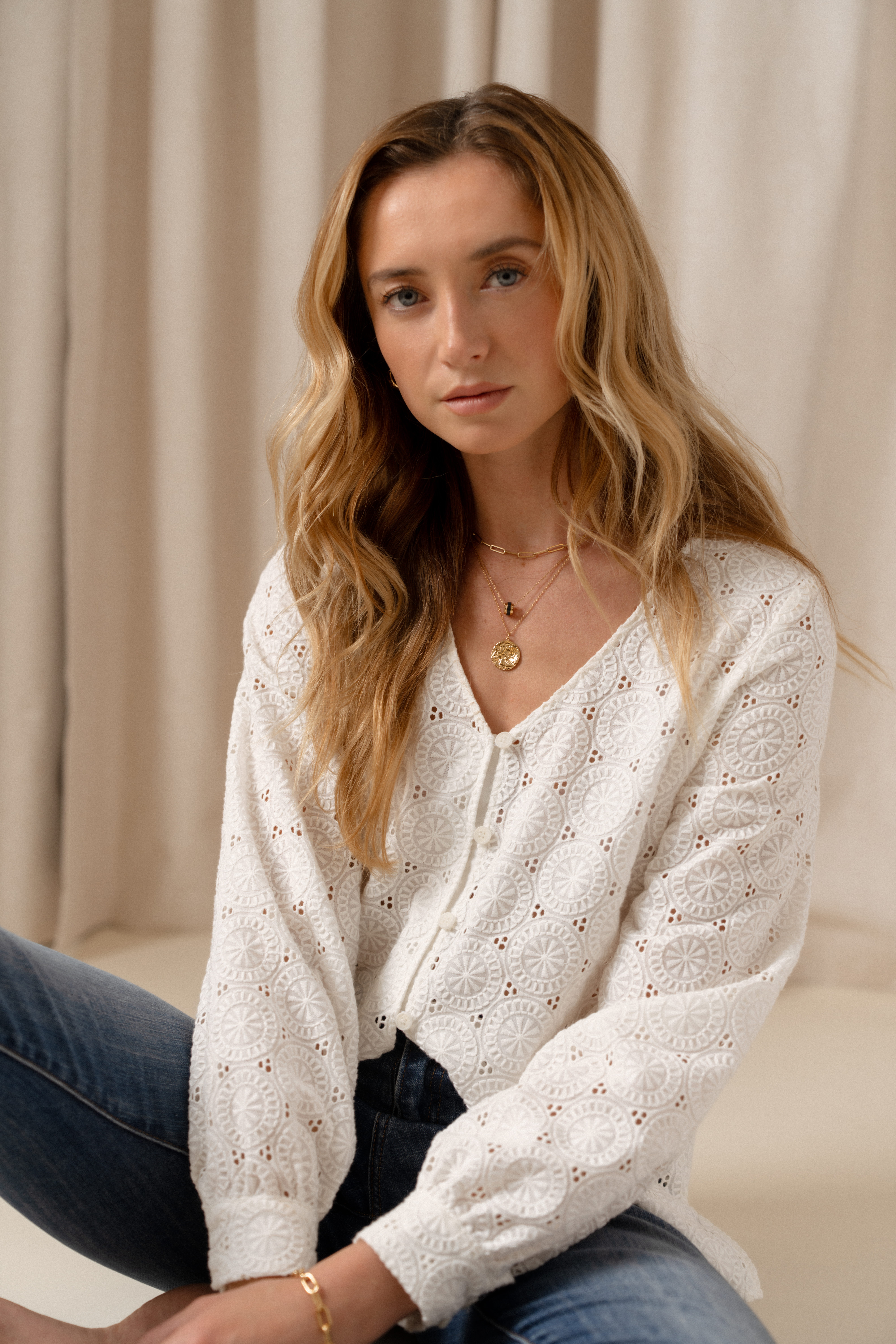 A model sitting on the ground in front of a beige curtain wearing a white button up blouse with a V neck line and a broderie anglaise texture.