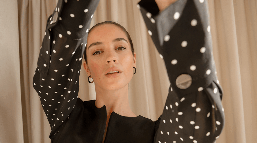 A model standing in front of a beige curtain holding her arms out toward the camera as she looks down at it. She is wearing a black blouse with black and white polka dot sleeves.