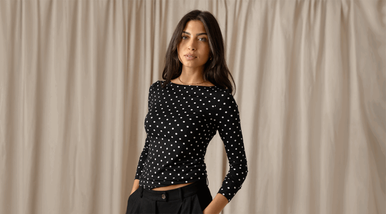 A model standing in front of a beige curtain wearing a black long sleeve top with a boat neckline and polka dot print. She stands with one hand tucked into her pocket.