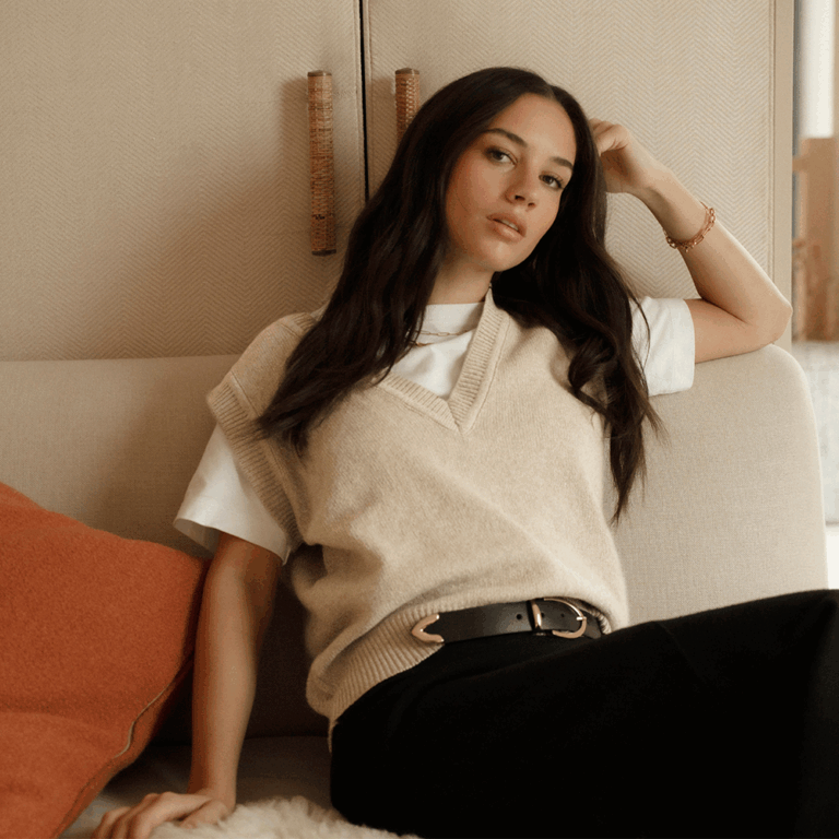 A model sitting on a beige sofa beside an orange pillow wearing a cream sweater vest over a white crew neck short sleeve t-shirt with wide leg black trousers