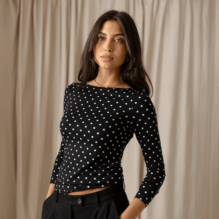 A model standing in front of a beige curtain wearing a black long sleeve top with a boat neckline and polka dot print. She stands with one hand tucked into her pocket.