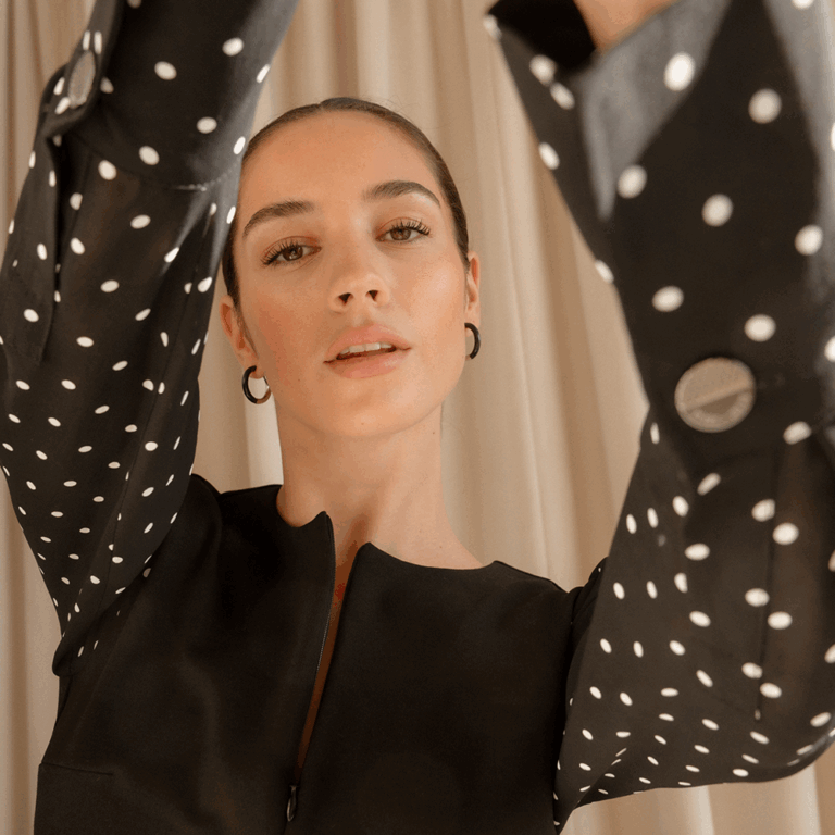 A model standing in front of a beige curtain holding her arms out toward the camera as she looks down at it. She is wearing a black blouse with black and white polka dot sleeves.