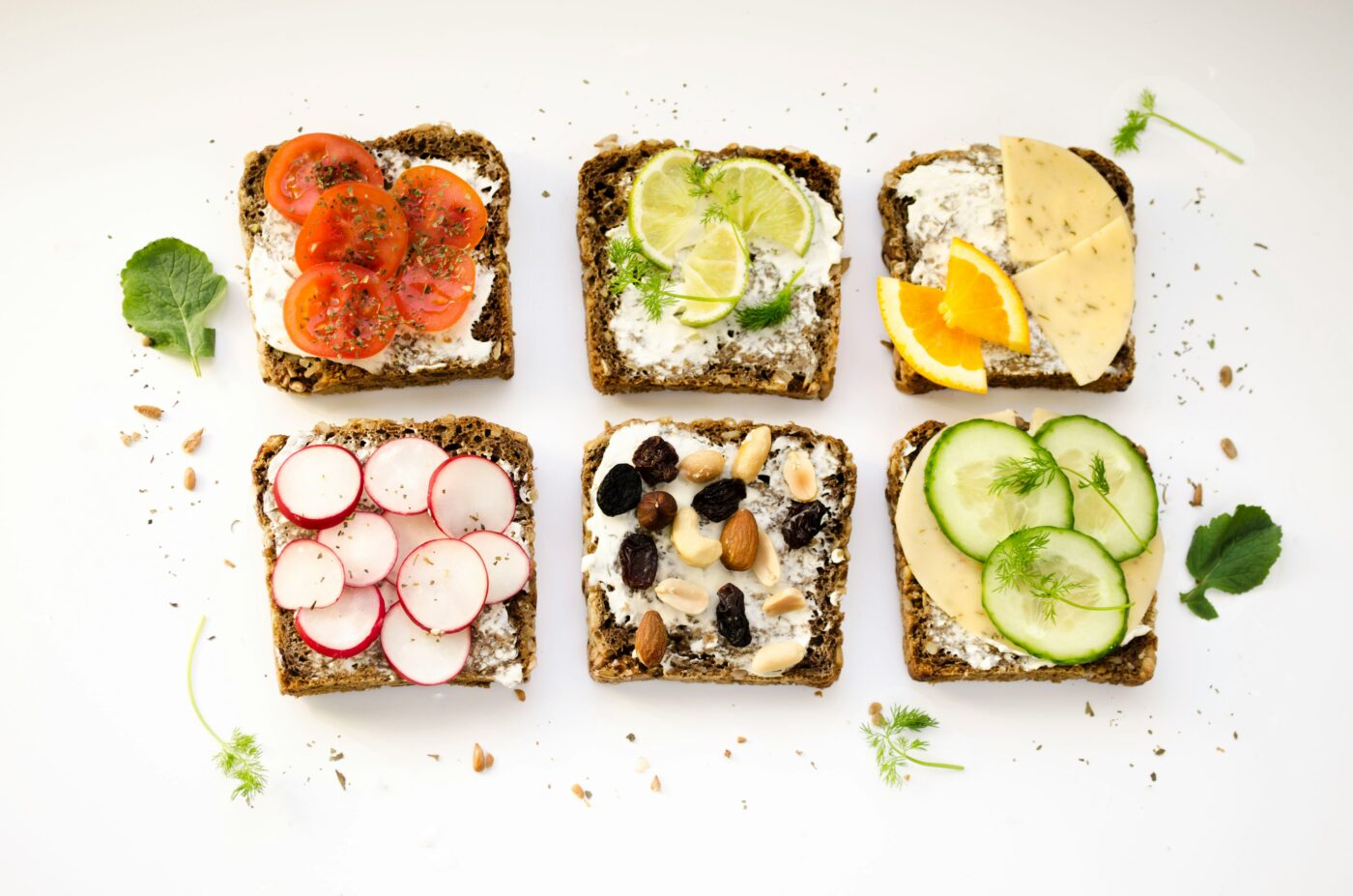 A selection of healthy toast options. One shows cream cheese with tomato, one shows cream cheese with lime, one shows cream cheese with slices of cheddar, one shows cream cheese with raddishes, one shows cream cheese with nuts, one shows cream cheese with cucumber.