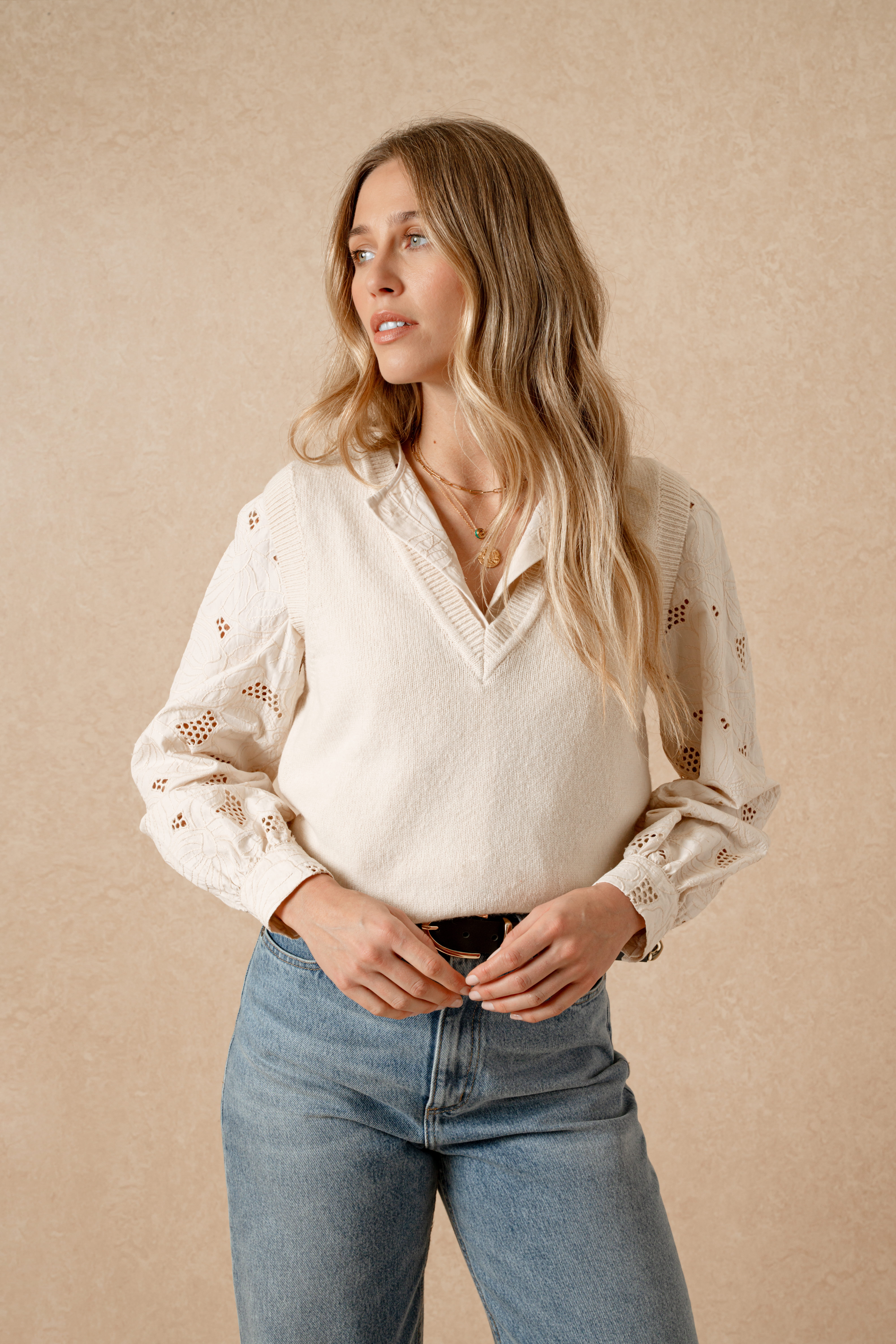 A model standing in front of a beige background wearing a cream broderie anglaise long-sleeved blouse underneath a V-neck sweater vest. It's tucked into a pair of mid-blue high-waisted jeans with a black belt.