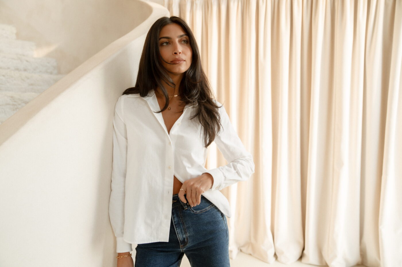 A model standing in front of a beige staircase, wearing a white button-up shirt and classic blue high waisted jeans.