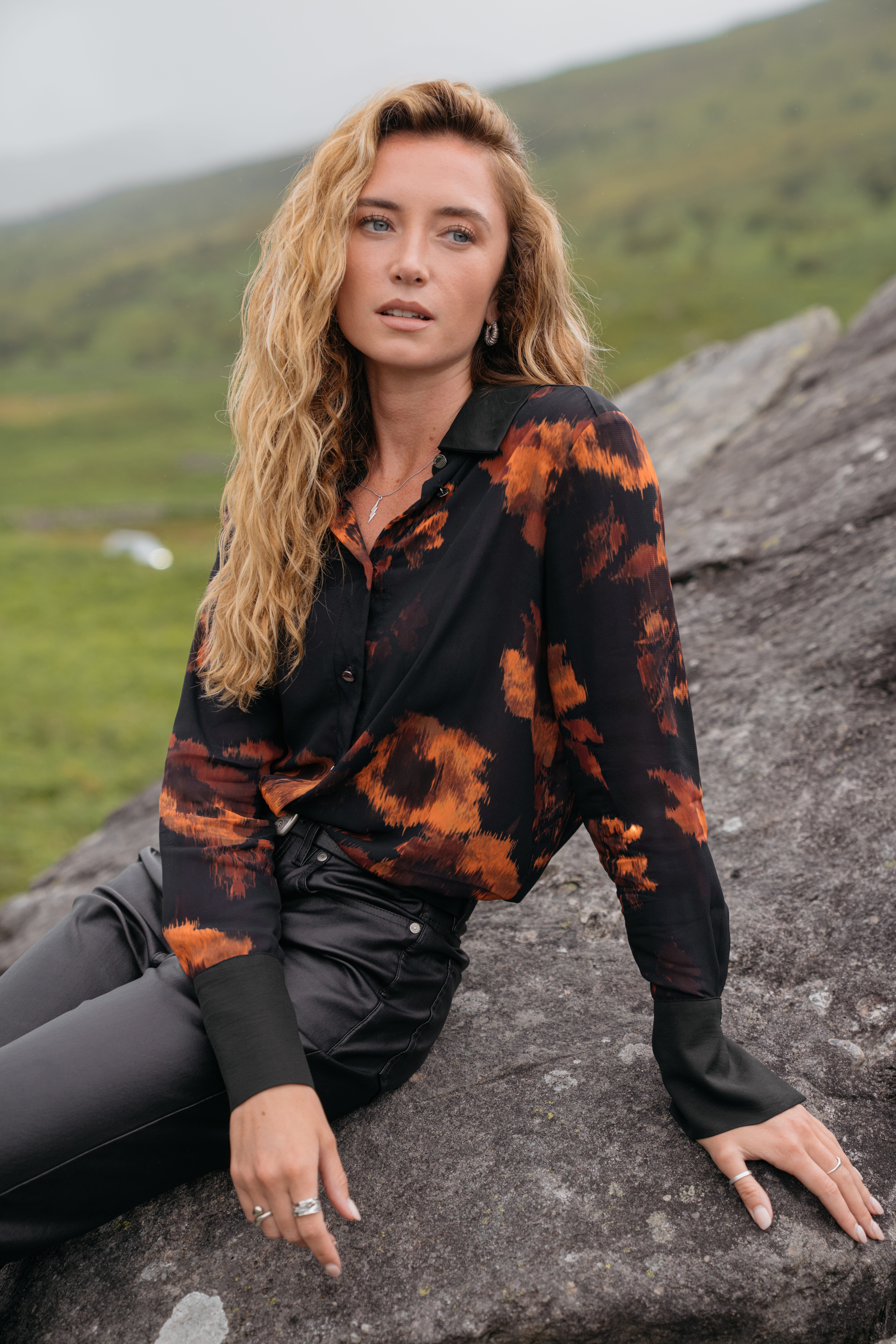 A model sitting on a rock wearing a pair of black leather jeans with a black long sleeved button-up shirt with orange flowers printed across the top.