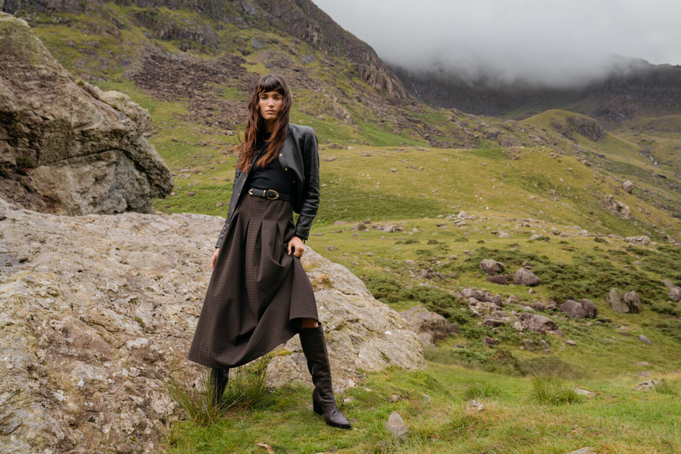 A model standing on a grassy landscape wearing a black leather jacket over a two-in-one style dress with a black long-sleeved bodice and checked midi skirt. It's styled with black leather boots. Each piece is from Saint + Sofia, a London-based fashion brand.