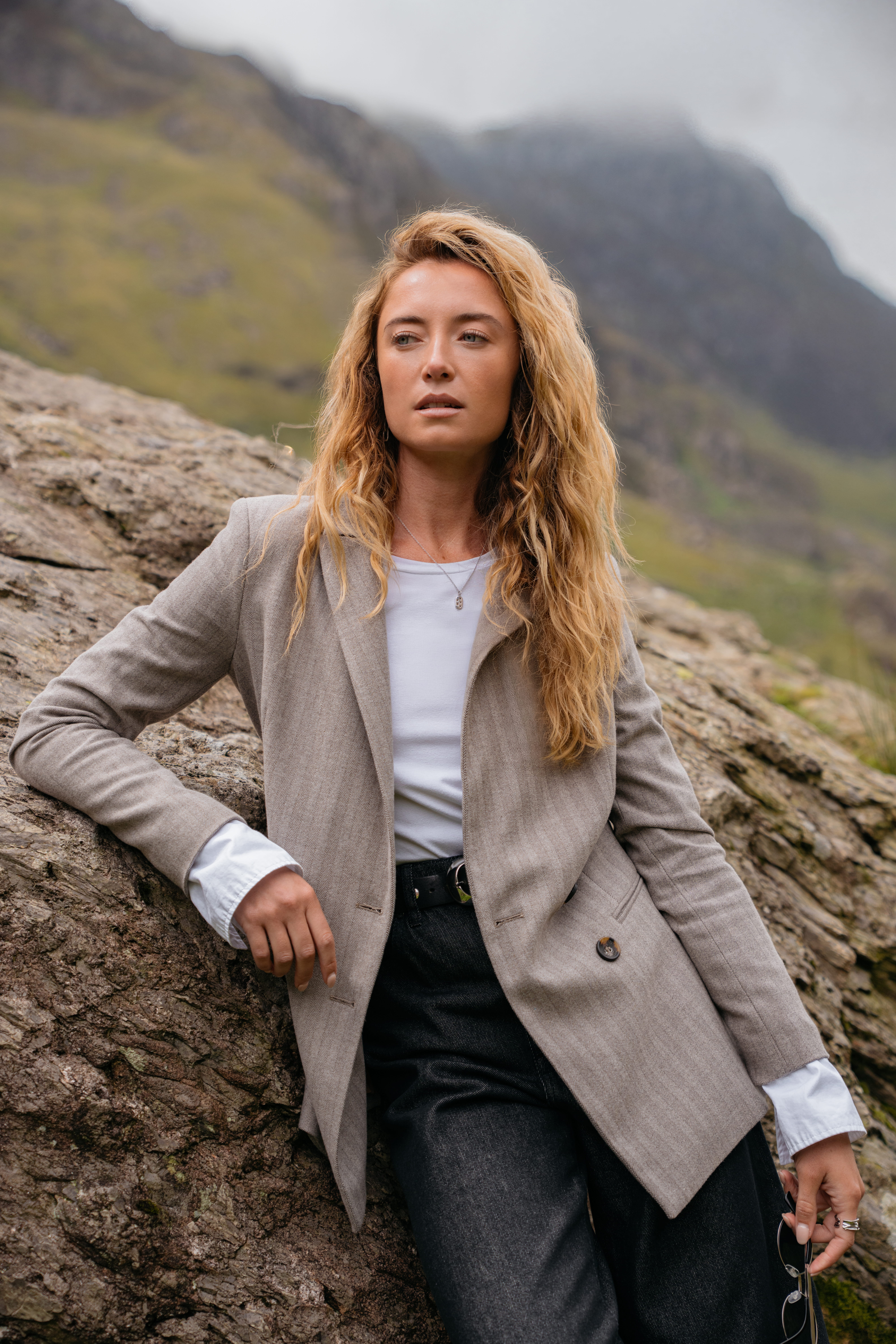 A model leaning against a rock wearing a grey blazer with a white long-sleeved shirt and a pair of black jeans.