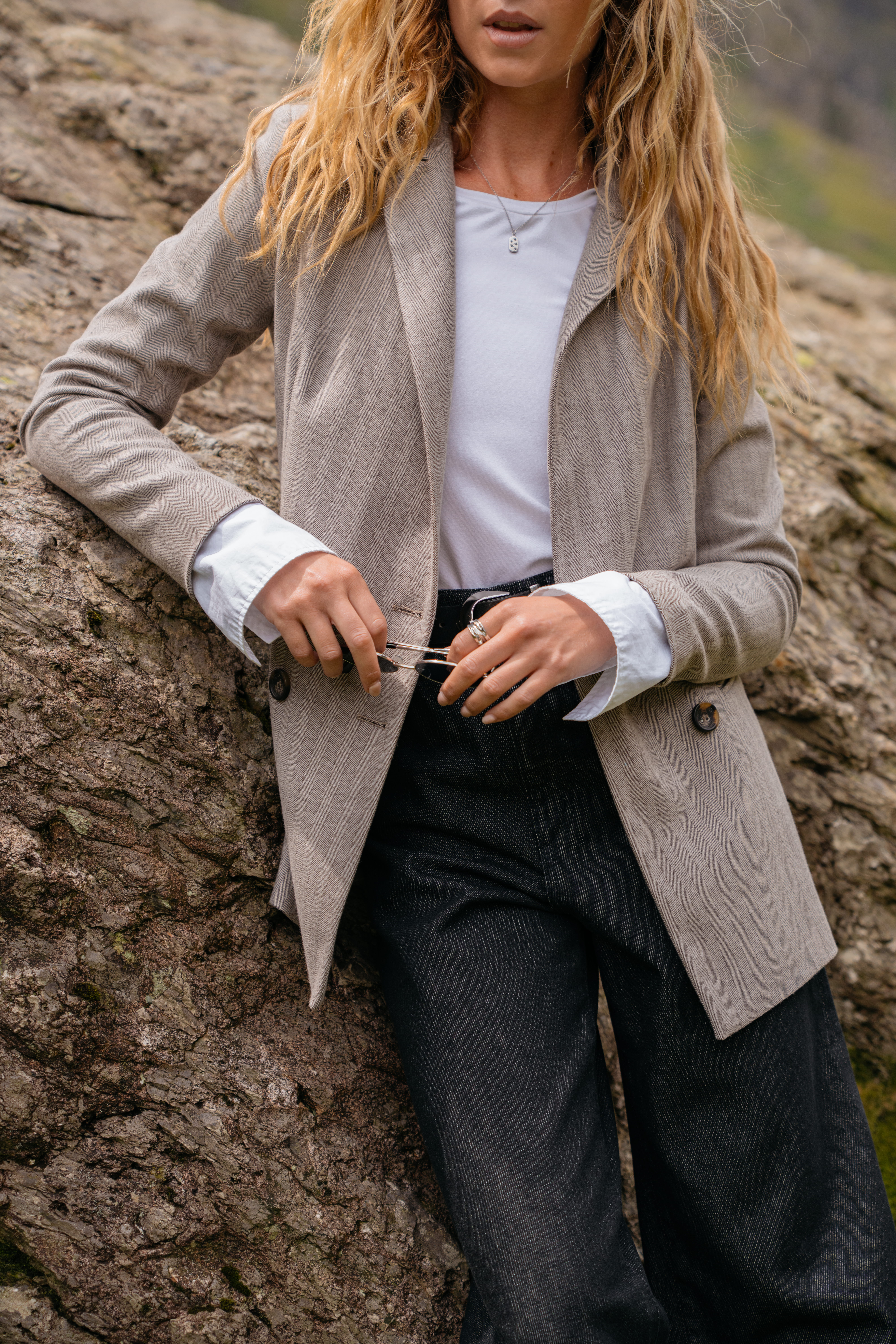 A model leaning against a rock wearing a grey blazer with a white long-sleeved shirt and a pair of black jeans.