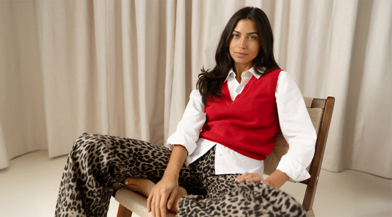 A model lounging in a wooden armchair against a beige curtain wearing a white button-up shirt under a red cashmere sweater vest with a V neckline and leopard print jeans