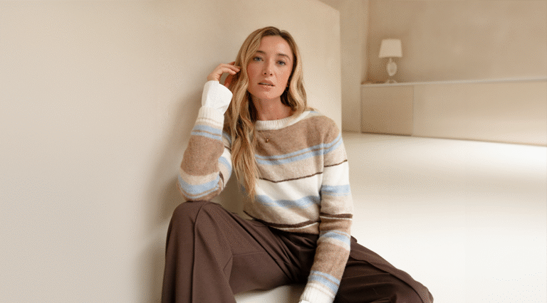 A model sitting on the ground in front of a beige background wearing a pair of brown wide leg trousers with a striped knitted jumper